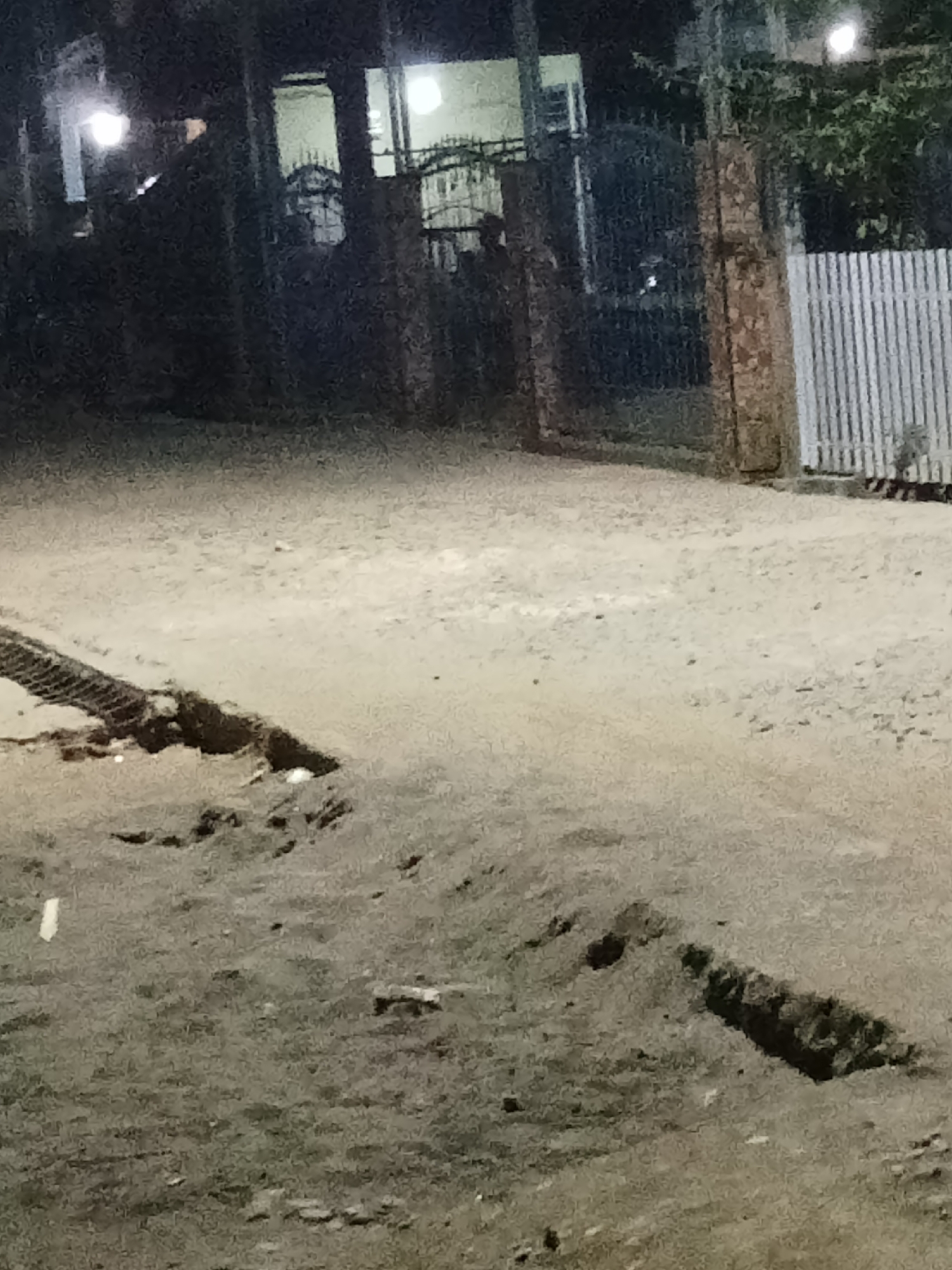 The image depicts an outdoor scene at night, likely in the city of Setia Jaya, Indonesia. The foreground is dominated by a dirt or gravel surface with several uneven patches and a drainage channel or trench. In the background, there appears to be a fence or gate structure, and beyond that, buildings with illuminated windows. The lighting suggests artificial light sources are present, possibly streetlights or lights from the buildings. The overall impression is that of a dimly lit outdoor area at night, possibly an alleyway, a construction site, or an unpaved road.