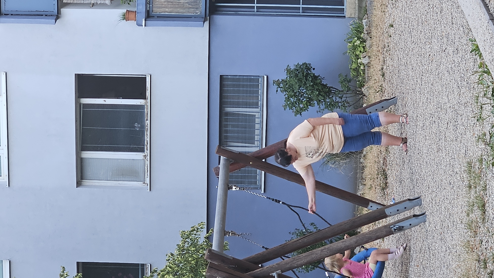 In a residential area of Bratislava, Slovakia, a woman in a peach-colored t-shirt and blue capri jeans stands near a wooden swing set. She is leaning forward, her right arm extended towards a swing where a young child in a pink top and shorts is seated. The woman appears to be interacting with the swing, possibly pushing it or retrieving something. The playground is covered in gravel, with sparse dry grass and a small bush nearby. Behind the playground, a light blue apartment building with several windows is visible. The windows have blinds, and one is slightly ajar. The scene suggests a typical outdoor activity for children and adults in a city setting.