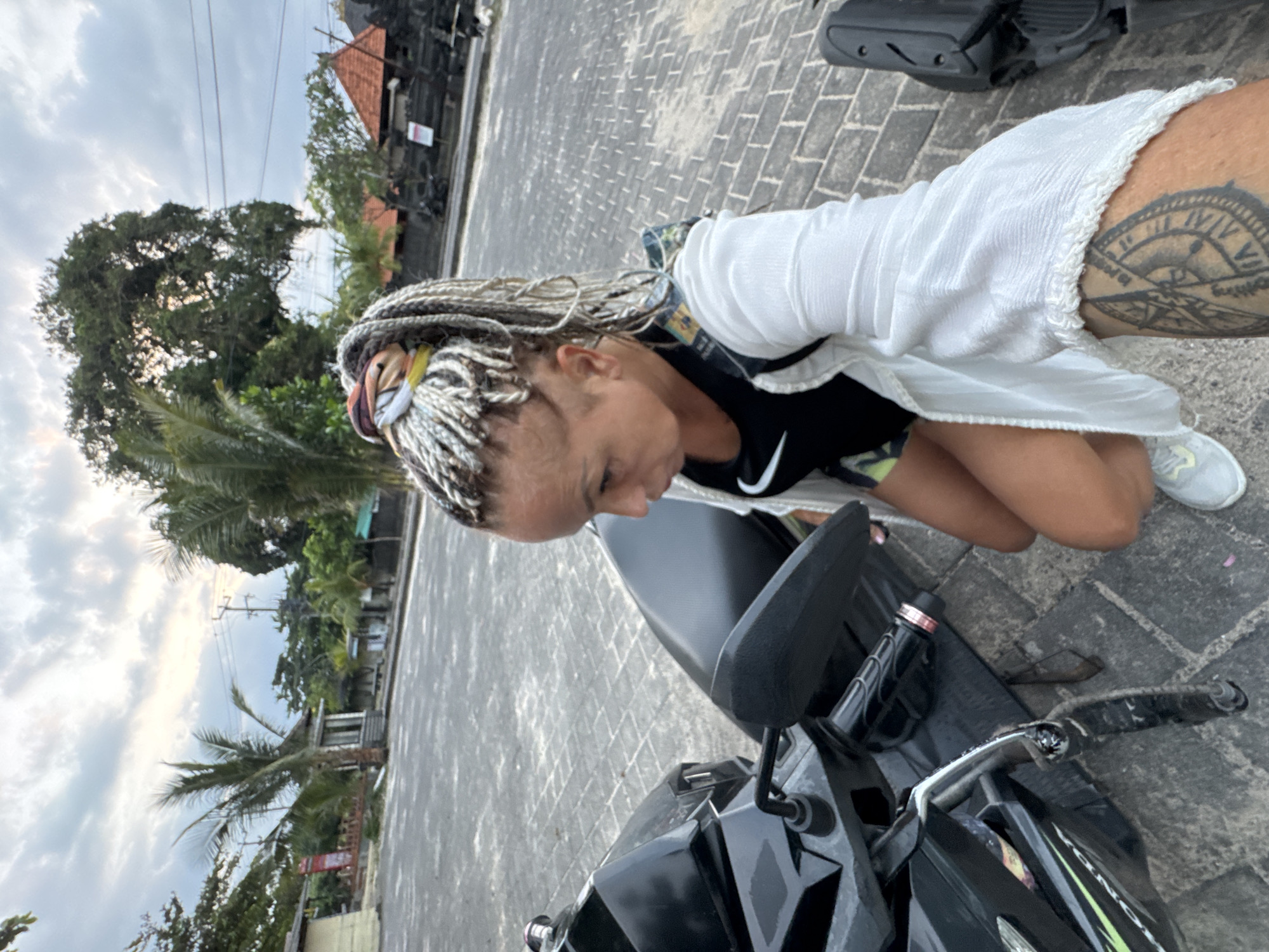 A medium shot captures a woman with blonde, braided hair, kneeling beside a black motorbike on a paved surface. She wears a white, fringed top over a black Nike tank top and light-colored sneakers. A tattoo is visible on her upper arm. The motorbike's handlebars, mirror, and part of the front are in the foreground. The background shows a street scene with buildings, trees (including palm trees), and a partly cloudy sky, suggestive of a location in Kuta, Indonesia. The woman appears to be looking down at the motorbike.