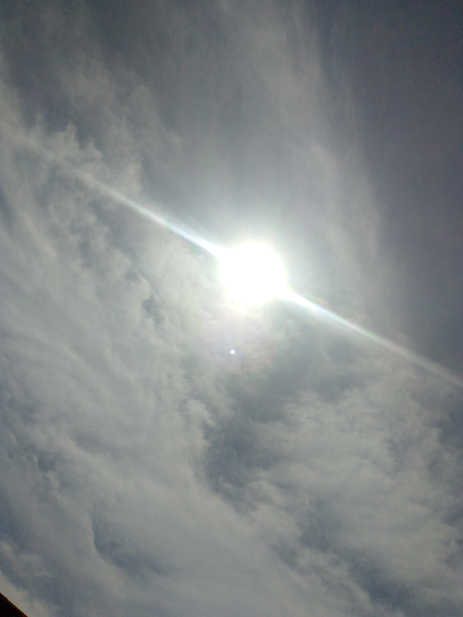 The image captures an overcast sky above Juan Griego, Venezuela. A bright, intense solar disc is positioned in the upper-left quadrant, partially obscured by cloud cover. A prominent, elongated sunbeam or lens flare emanates diagonally from the solar disc, traversing the central portion of the frame. The sky is filled with a mixture of grey and white clouds, exhibiting various densities and textures, consistent with a partly cloudy or altostratus formation. A smaller, distinct white circular artifact, likely a lens flare, is visible below and to the left of the main solar presence. A dark, indistinct edge of a structure is minimally visible in the extreme bottom-left corner, indicating an upward-facing perspective from ground level. No human subjects or their direct interactions are present.