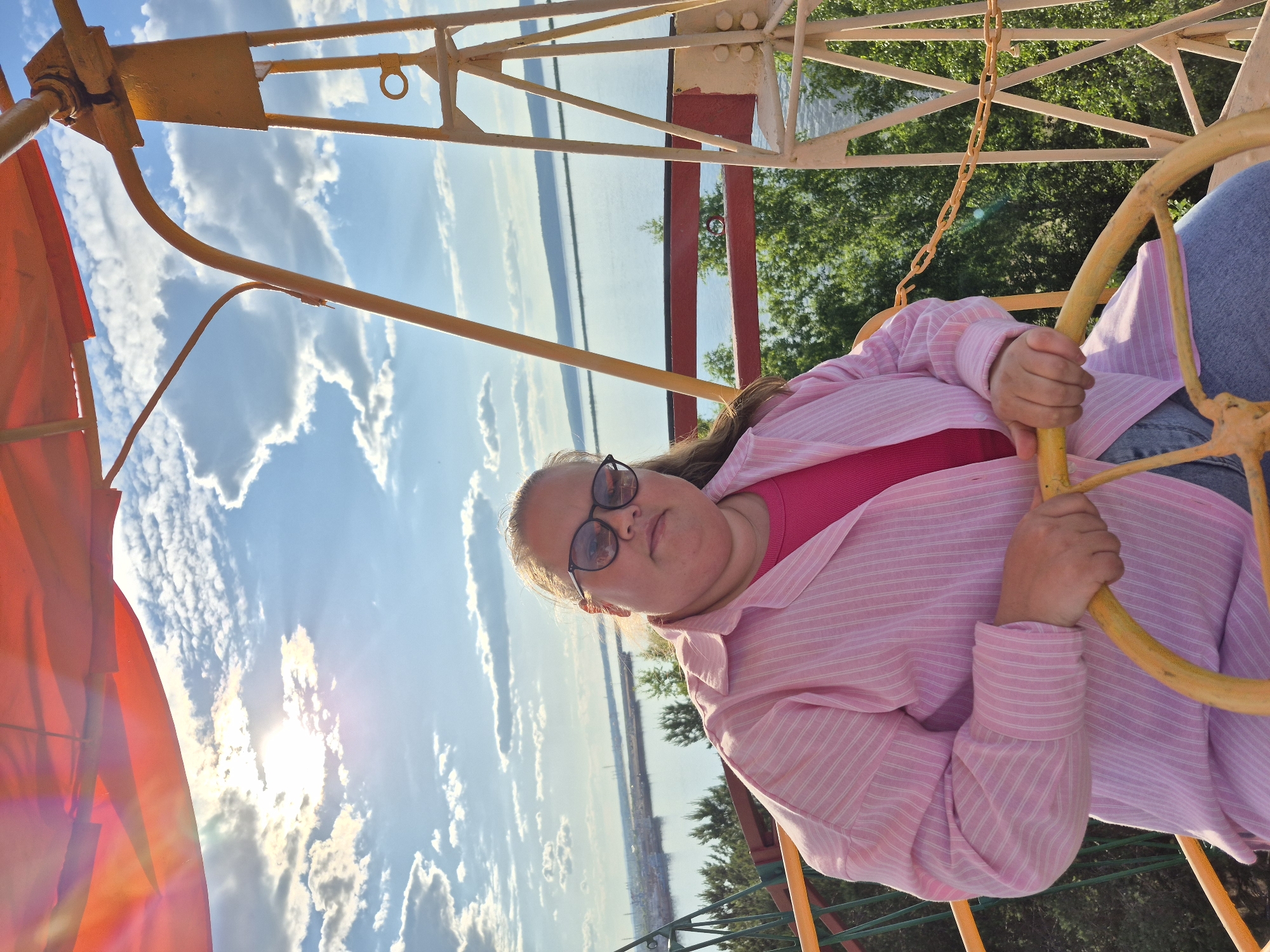 The image displays a Caucasian woman situated within a metal observation structure, likely a Ferris wheel gondola or viewing platform, in Chaikovsky, Russia. The woman, appearing to be in her late 20s to 40s, has light brown hair and wears dark-framed eyeglasses. She is dressed in a pink and white striped long-sleeved button-down shirt over a solid fuchsia top, paired with light blue denim jeans. Her hands are positioned on a circular, worn yellow metal handle or wheel directly in front of her, and she looks directly at the camera with a neutral expression.
An orange fabric canopy is visible above her, partially obscuring the sun, which is intensely bright in the upper left portion of the sky, casting significant lens flare. The sky is clear blue with numerous scattered white cumulus clouds. The surrounding structure is made of yellow metal, featuring vertical and diagonal supports, a red horizontal bar visible below the woman, and a segment of a yellow chain on the right.
Below the structure, a dense expanse of green deciduous trees is visible. In the background, a large body of water, identified as the Votkinsk Reservoir, stretches across the horizon. A distant landmass, including a forested shoreline and what appears to be a port or industrial area with visible vessels or structures, is discernable on the far bank of the reservoir towards the left.