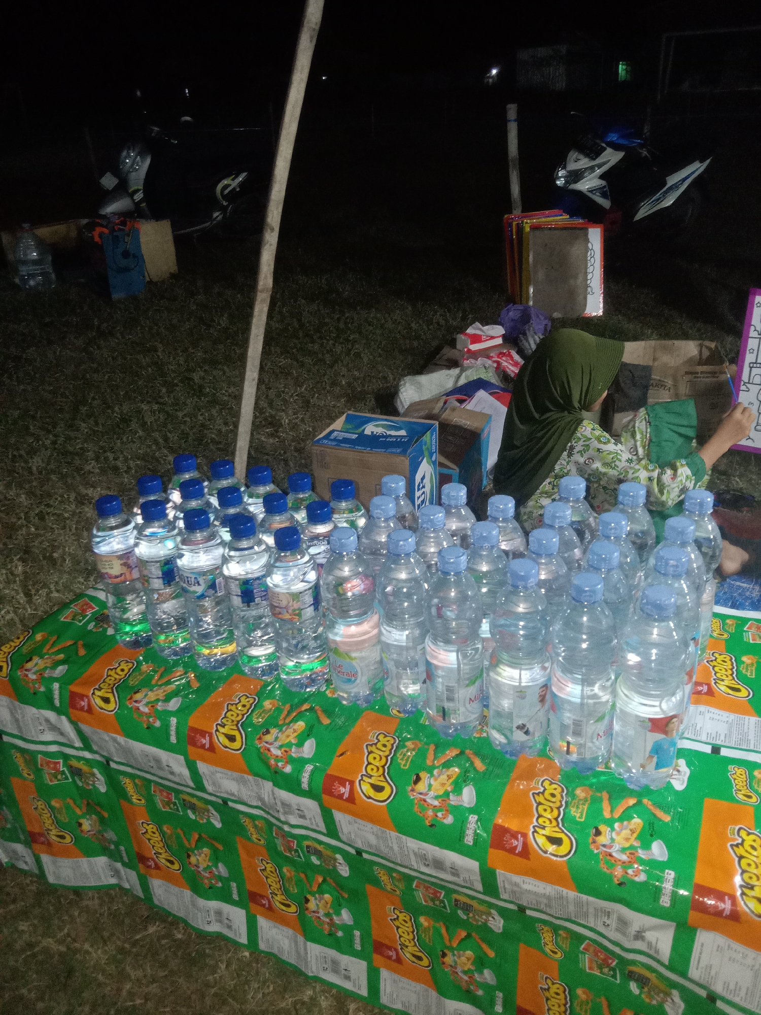 In the city of Jumput, Indonesia, at night, a collection of bottled water and snacks is displayed on a table covered with a Cheetos-branded tablecloth. The table is positioned on a patch of grass. In the background, two motorcycles are parked, and several boxes and bags are scattered around. Two individuals are visible; one is wearing a green headscarf and a patterned shirt, and appears to be drawing or writing on a board. Another person, also wearing a green headscarf, is partially visible next to them. The lighting is dim, with some artificial light sources illuminating the scene.