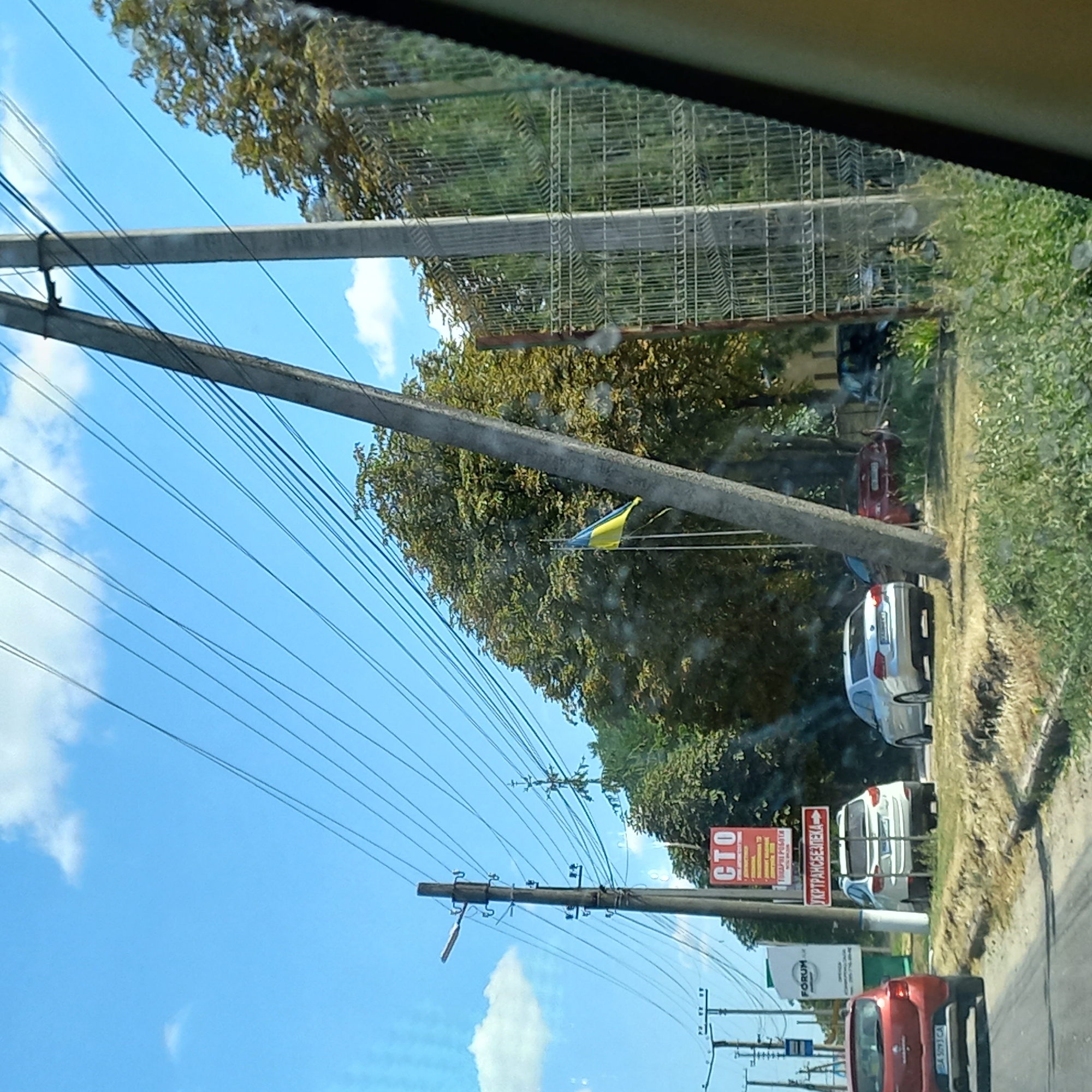 The image shows a street scene in Kropyvnytskyi, Ukraine. A road is visible in the foreground, with a red car on the left and a white SUV and silver sedan parked on the right. A tall utility pole with numerous wires runs diagonally across the frame, with a Ukrainian flag attached to it. Several other utility poles and overhead wires are also present. In the background, there are trees and a wire fence. A sign that says "СТО" in red letters is attached to a utility pole. The sky is blue with some scattered clouds. The overall impression is of a typical urban or suburban street on a clear day.