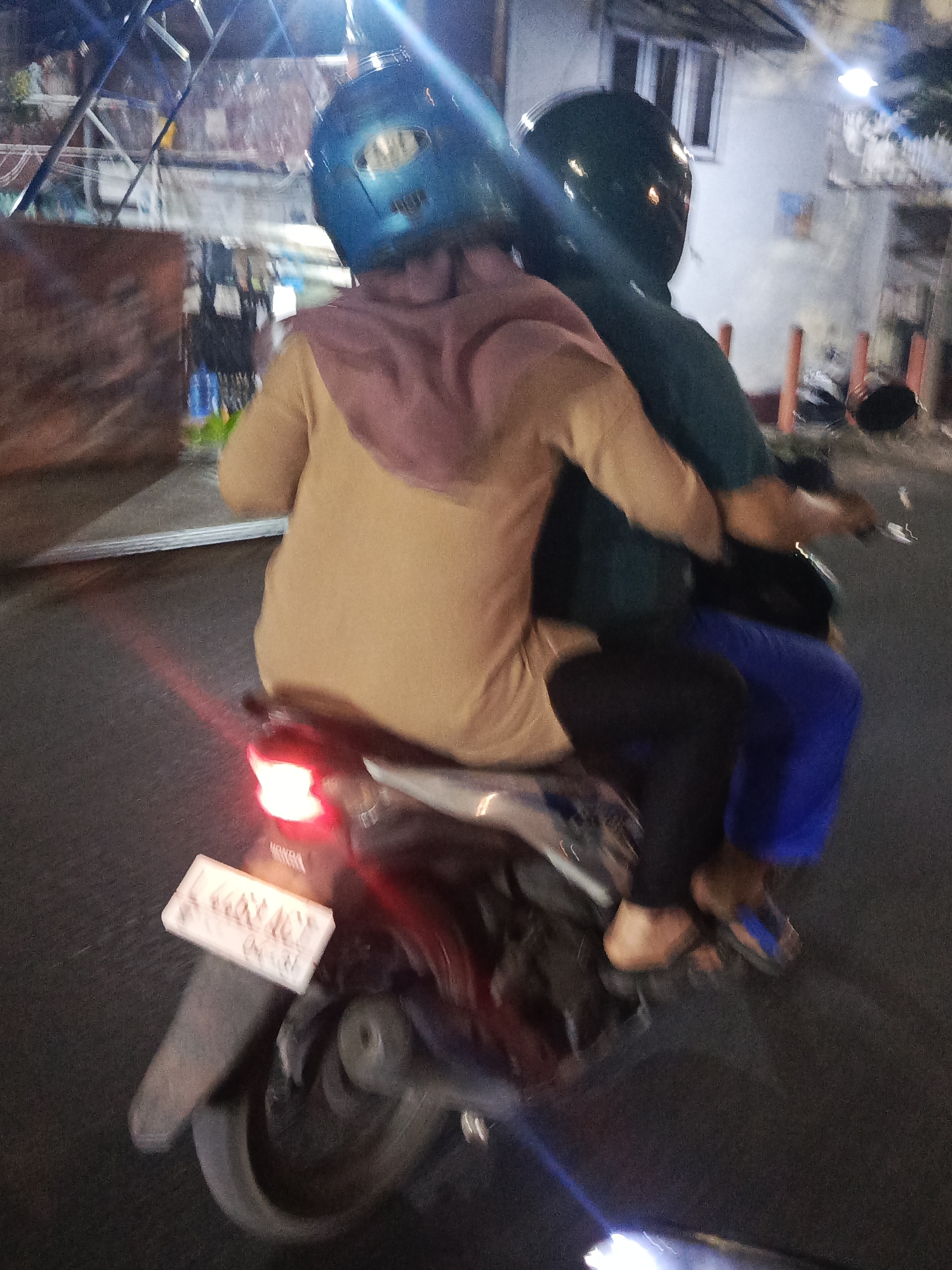 Two individuals are riding a motorcycle at night on a city street in Surabaya, Indonesia. The person in front is wearing a blue helmet and a tan-colored shirt with a purple headscarf. The person behind is wearing a dark green helmet and a dark green shirt. Both are wearing dark pants. The motorcycle has a red taillight and a license plate that is partially visible. The street appears to be asphalt, and there are buildings and structures in the background, suggesting an urban environment. The lighting is dim, indicative of nighttime, with some bright lights from artificial sources contributing to the scene's illumination. The overall image quality is low, with motion blur present due to the movement of the motorcycle.