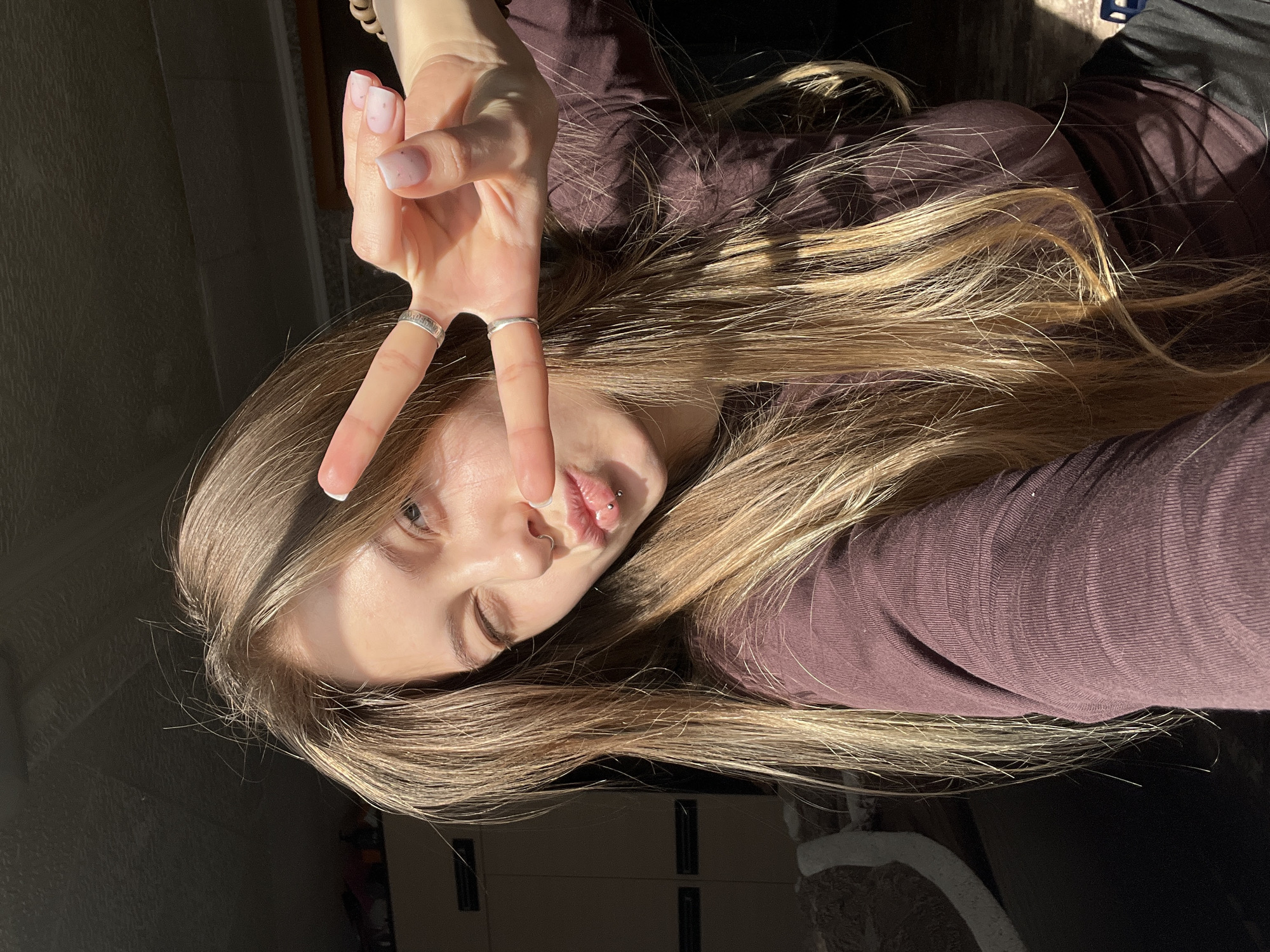 A person, likely a young woman, is posing for a selfie. She is lying down with her head tilted towards the camera, making a peace sign with her left hand. She has long blonde hair and is wearing a purple long-sleeved shirt. She has piercings in her nose and lip and is wearing two rings on her fingers. The lighting is uneven, with the upper left portion of the image brightly lit and the lower portion in shadow. The background is indistinct but appears to be an interior setting. There is no definitive information to place this photo in Фоки, Russia.