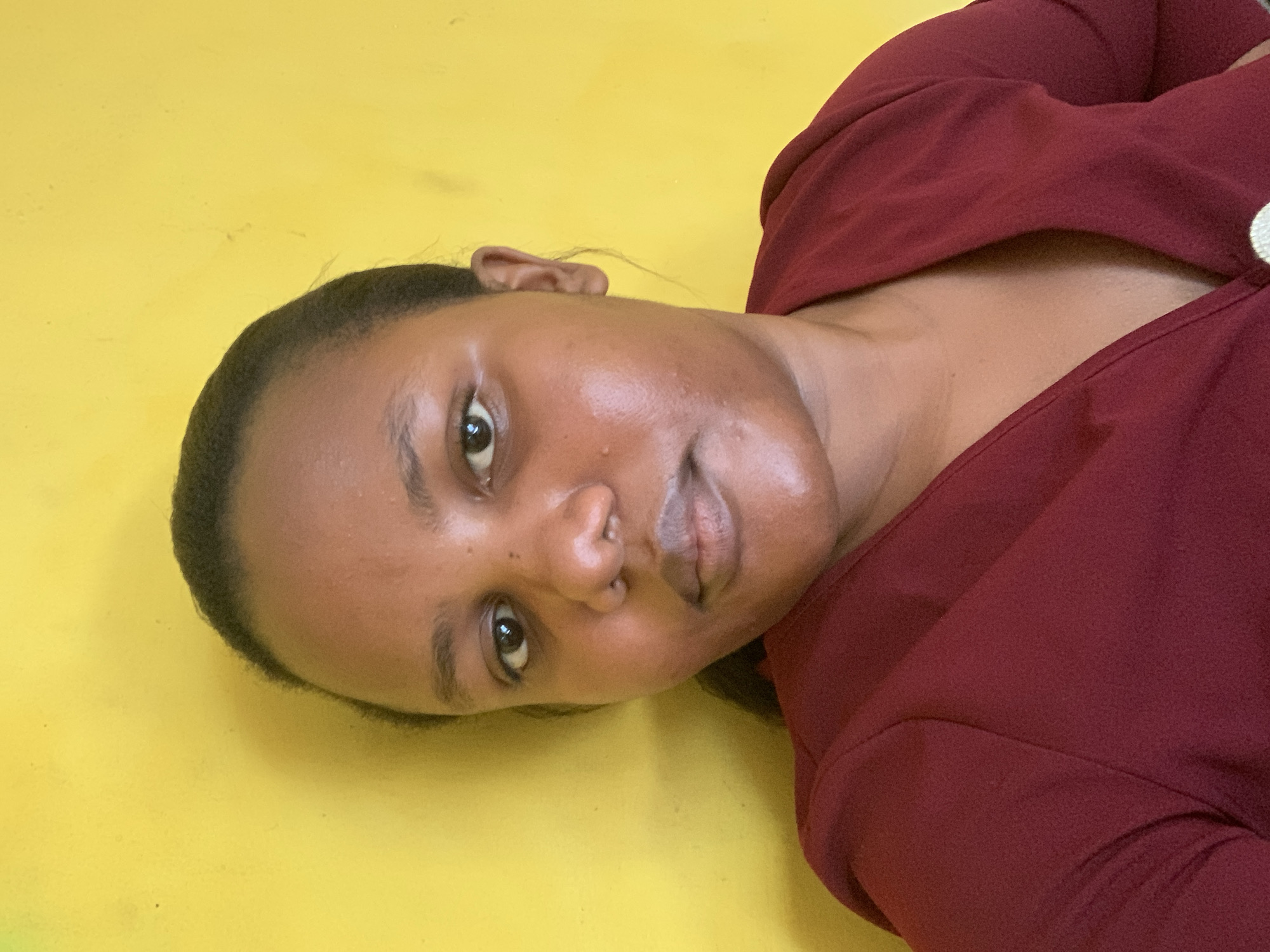 A medium shot depicts a dark-skinned woman with dark hair, positioned on a yellow background, wearing a maroon top. No elements point to a specific location in Dar es-Salaam, Tanzania. There are no other people, objects, or actions visible in the scene.
