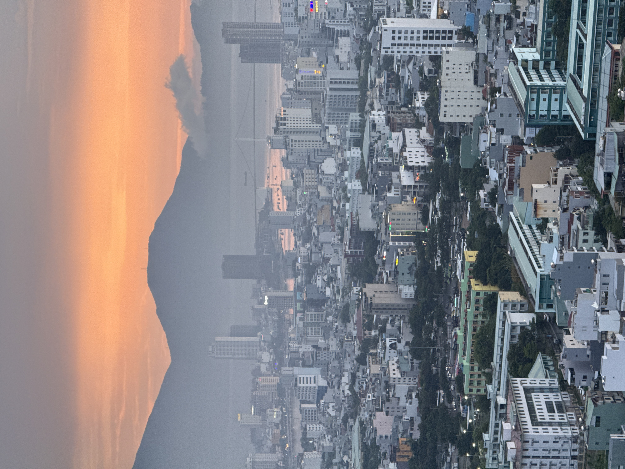 This is a high-angle, vertical, full-frame photograph of the city of Da Nang, Vietnam, during what appears to be sunrise or sunset, given the orange and pink hues in the sky. The foreground and midground are dominated by a dense urban landscape with a variety of buildings, ranging from multi-story apartment complexes and office buildings to smaller residential structures. Architectural styles vary, with many buildings featuring light-colored facades, some with prominent window patterns, and others with balconies. Greenery, such as trees and small parks, is interspersed throughout the city.
In the background, a body of water, likely the Han River, is visible, reflecting the colors of the sky. A suspension bridge, characteristic of Da Nang's modern infrastructure, spans the water. Beyond the river and the bridge, mist-shrouded mountains rise, framing the city. The sky transitions from a vibrant orange near the horizon to a muted gray at the top of the frame, with a few wispy clouds present. The overall impression is one of a sprawling coastal city at the edge of a natural landscape, illuminated by the soft light of dawn or dusk. No people are visible in the image.