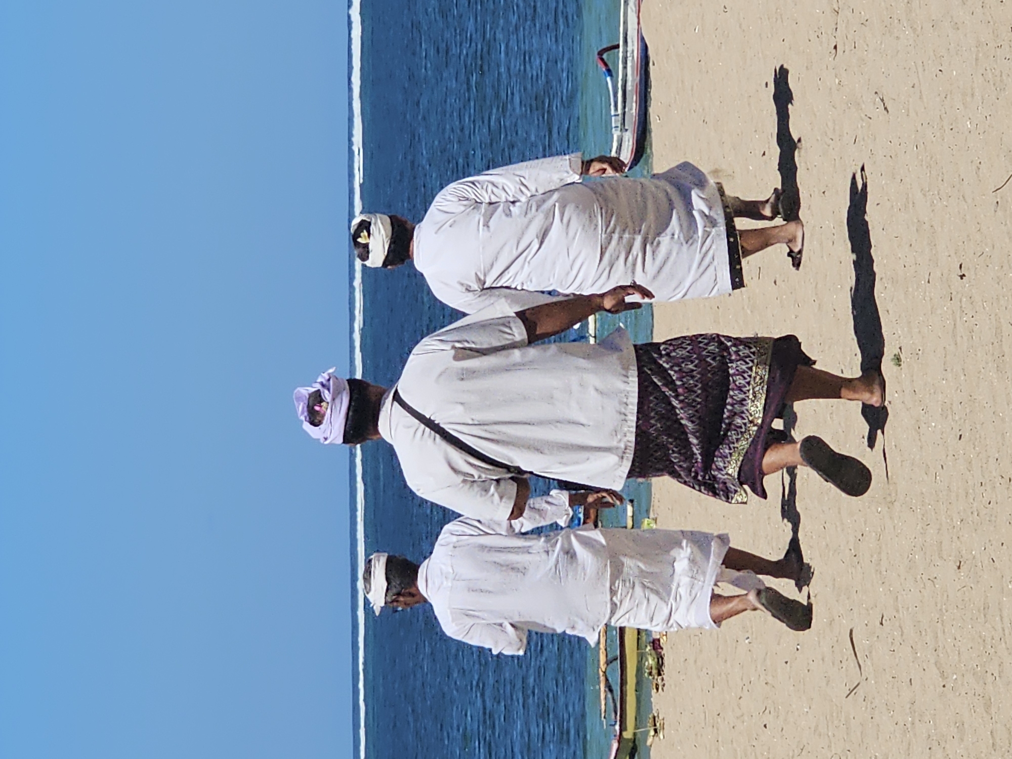Three men in traditional Balinese attire are walking away from the viewer on a sandy beach. The man in the center wears a purple headdress, a white collared shirt, and a patterned sarong. The man to his left wears a white cap, a white long-sleeved shirt, and a white sarong. The man to his right wears a white headwrap with a flower, a white long-sleeved robe, and a white sarong. All three men are wearing sandals. They are walking towards the ocean, which is a deep blue with a white wave line in the distance. The beach is light brown sand with some debris scattered on it, including what appears to be parts of a boat in the background. The sky is clear and blue. The scene appears to be in Denpasar, Indonesia, given the traditional attire and the coastal setting.