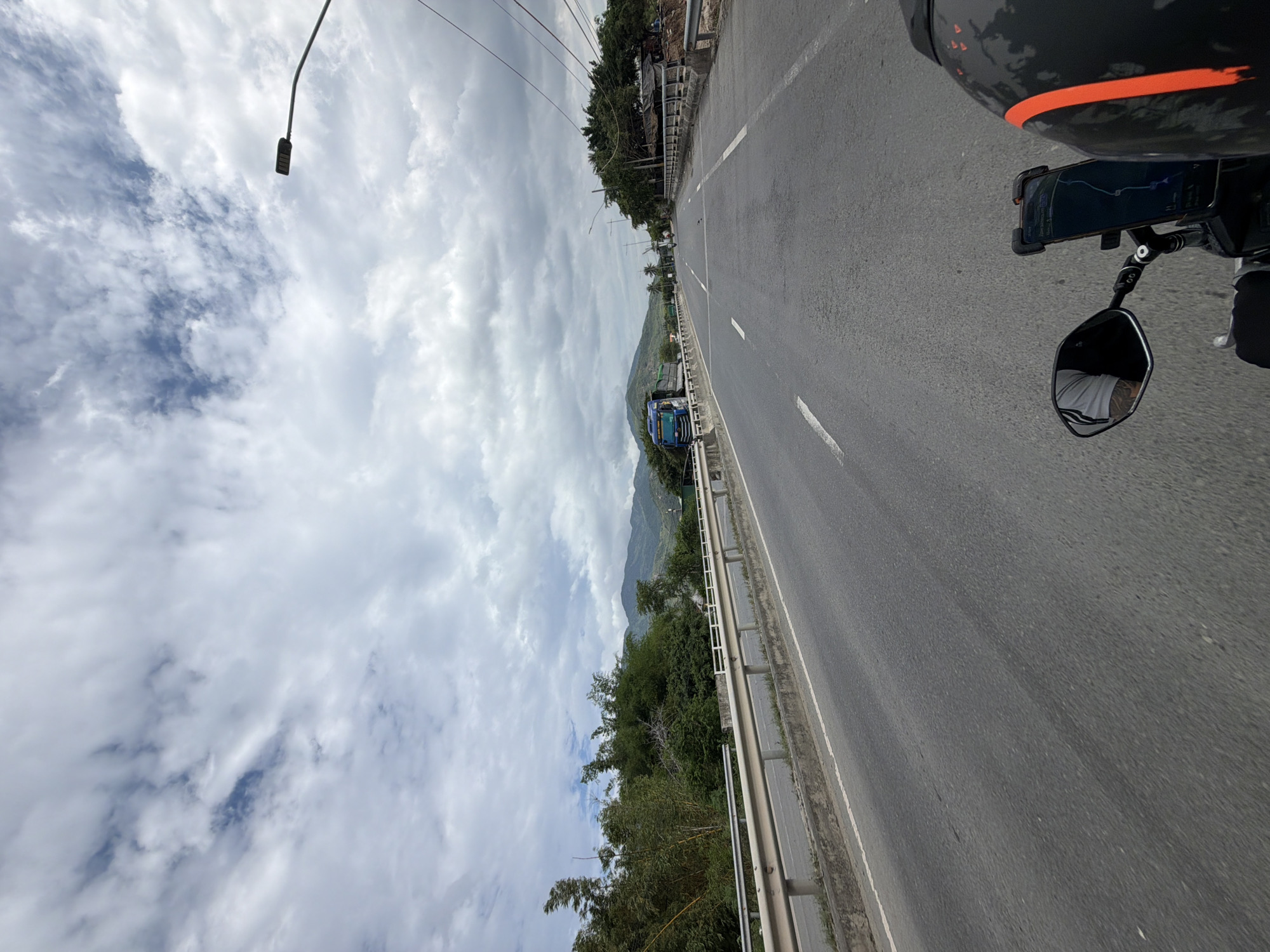 The image captures a first-person perspective from a motorcycle traveling on a road, as if viewed from the rider's right side. In the immediate foreground on the right, parts of the motorcycle are visible, including a black helmet with an orange stripe, a handlebar, and a smartphone mounted to the handlebar displaying a map application. A rearview mirror reflects a portion of the rider's torso and arm, clad in a white t-shirt.
The asphalt road, marked with white dividing lines, extends into the distance. A metal guardrail parallels the left edge of the road. Beyond the guardrail, a lush, green hillside covered with dense trees and vegetation descends. A blue truck is visible in the mid-distance on the road, partially obscured by the guardrail. Further in the background, a range of green mountains dominates the horizon.
The sky is heavily overcast with numerous white and gray clouds, though some patches of blue sky are visible. A street lamp, partially cut off, hangs from a pole in the upper left corner. The scene depicts a daytime journey through a scenic, mountainous, and forested landscape, indicative of a location like Cam Ranh Ward, Vietnam. The weather appears bright despite the cloud cover. No specific text is legible beyond the graphical interface of the navigation app.