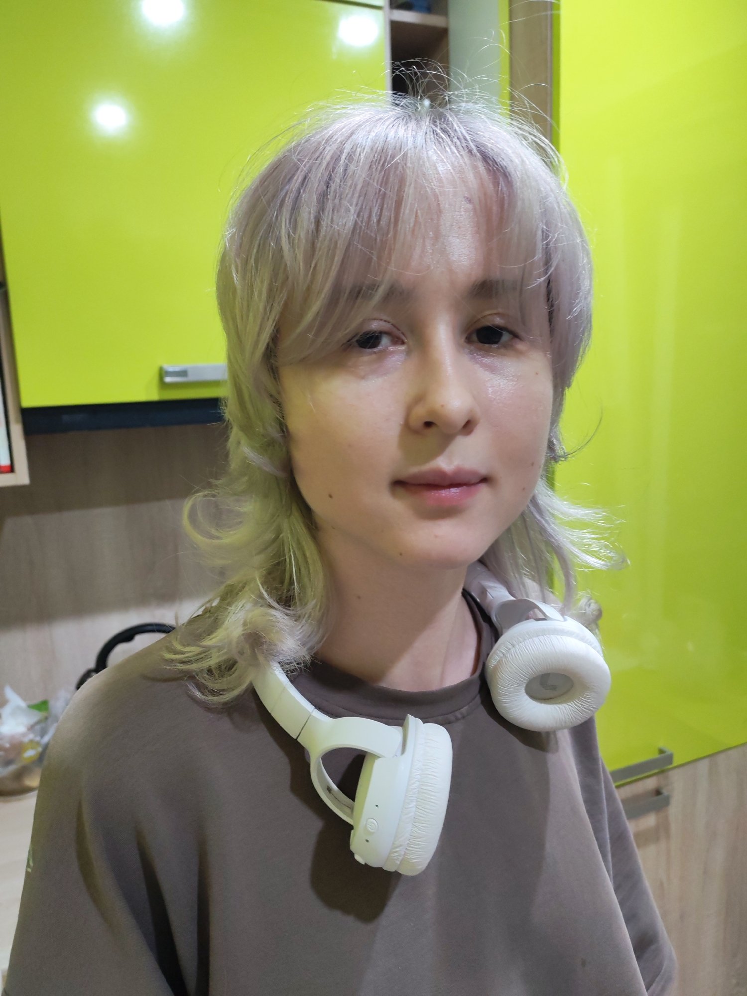 A person with light blonde, choppy hair is shown from the chest up, wearing a brown sweatshirt and white headphones around their neck. They have fair skin, dark eyes, and light pink lips. The background features a bright lime green cabinet and a wooden surface with some items visible on the left. The scene appears to be indoors, possibly a kitchen, with artificial lighting. There is no discernible indication of the city of Chaikovsky, Russia within the image.