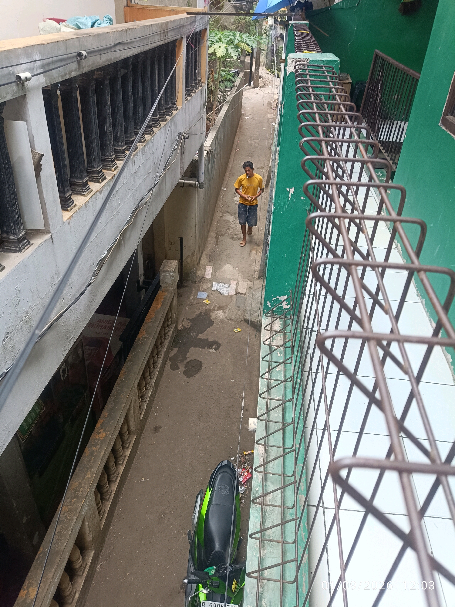 A man wearing a yellow t-shirt and blue shorts is walking down a narrow alley. He is holding a cigarette in his right hand. The alley is lined with buildings on both sides. The building on the left has a stone railing with black pillars. The building on the right is painted green and has a metal grate in front of it. There is a motorcycle parked in the alley on the right side. The ground is made of asphalt and is dirty. There are some pieces of trash on the ground. A timestamp in the lower right corner indicates the date 01/09/2026 and time 12:03.
