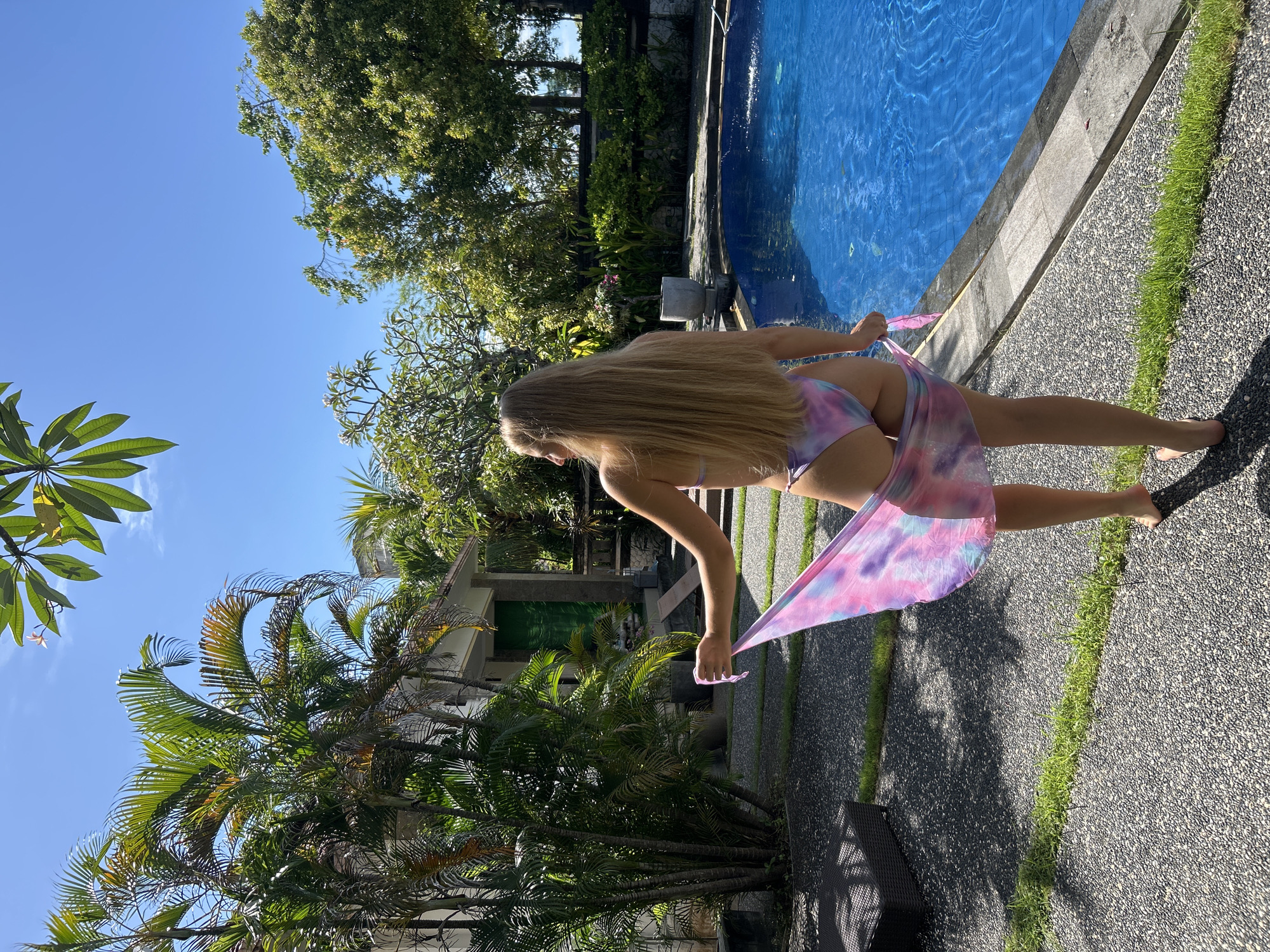 The image depicts a woman in a bikini standing next to a swimming pool. She is wearing a tie-dye sarong. The background includes tropical vegetation, a building, and a clear blue sky. The location is Kuta Selatan, Indonesia.