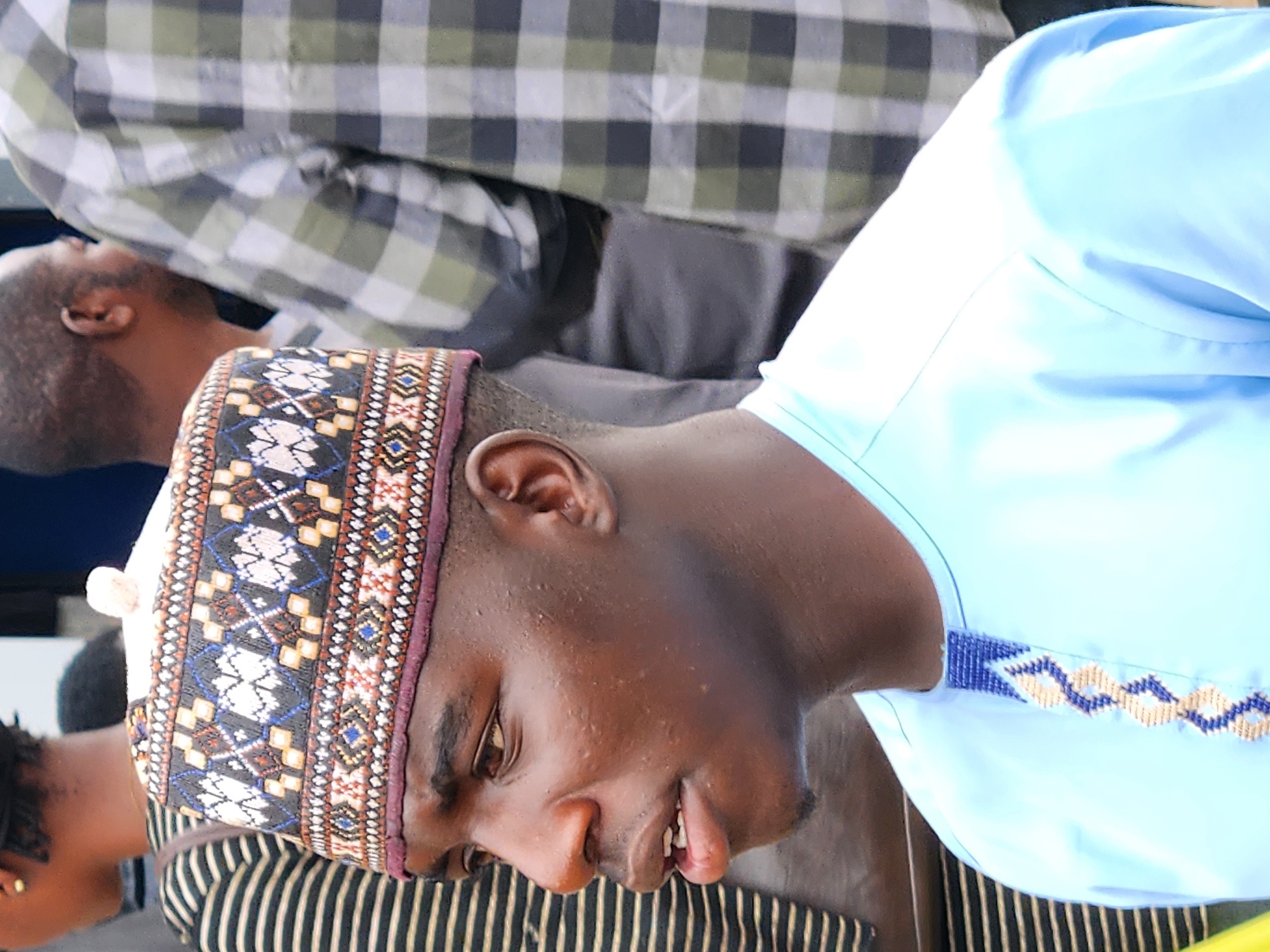 A close-up portrait of a young Black man wearing a light blue traditional garment and a patterned cap. The man is looking to his left. The background is blurred, showing other people and plaid shirts. The setting appears to be outdoors, possibly in a city.