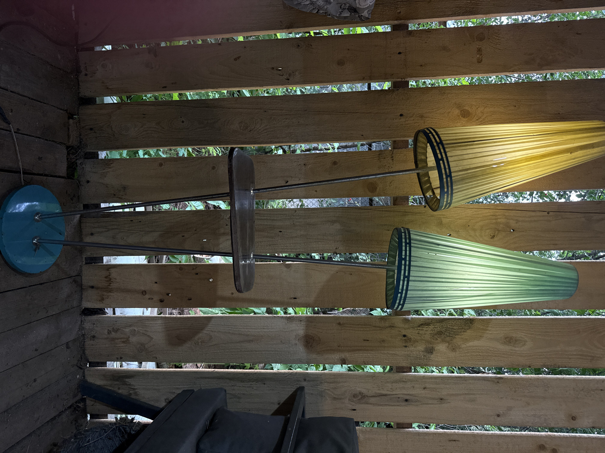 A tall, double-headed floor lamp with pleated shades, one yellow and one green, stands on a wooden deck in front of a vertical wooden plank fence. The fence is weathered and has gaps between the planks, revealing green foliage behind it. The lamp has a dark brown, almost black, tiered shelf supported by thin metal poles. The base of the lamp is a flat, teal-colored circle. To the right of the lamp, a dark gray cushioned chair or sofa is partially visible, along with some coiled black material. The lighting suggests an outdoor evening or shaded area. The location is identified as Khokhryaki, Russia.