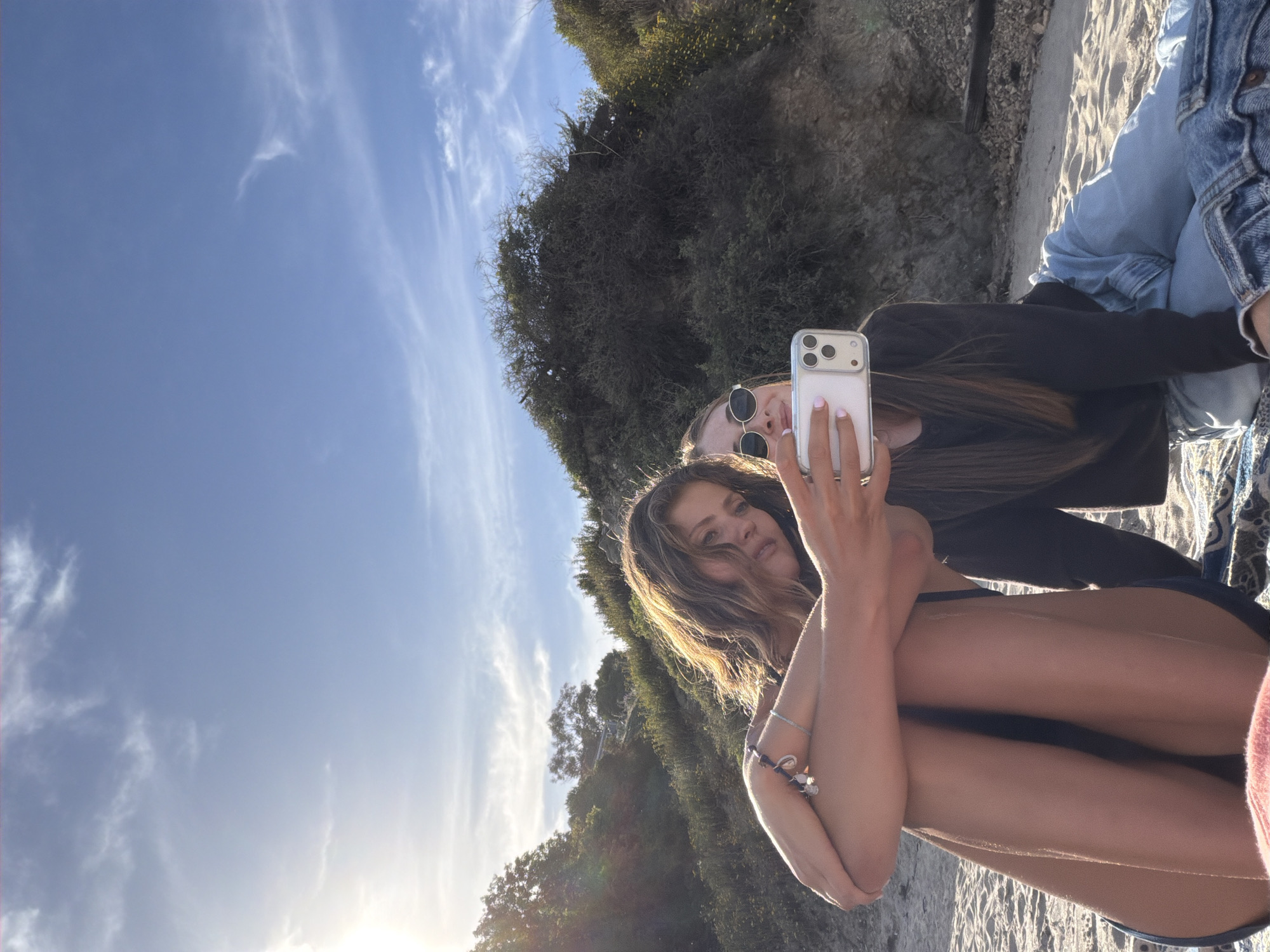 Two young women are posing for a selfie on a beach in Malibu, California. One woman is holding a smartphone, a silver-colored iPhone, with her left hand extended. She is wearing a dark-colored swimsuit. The second woman is behind her, wearing a long-sleeved top and sunglasses. The beach has light-colored sand, and behind them is a steep bank covered in vegetation. The sky is light blue with wispy clouds. The sun is visible, causing lens flare.