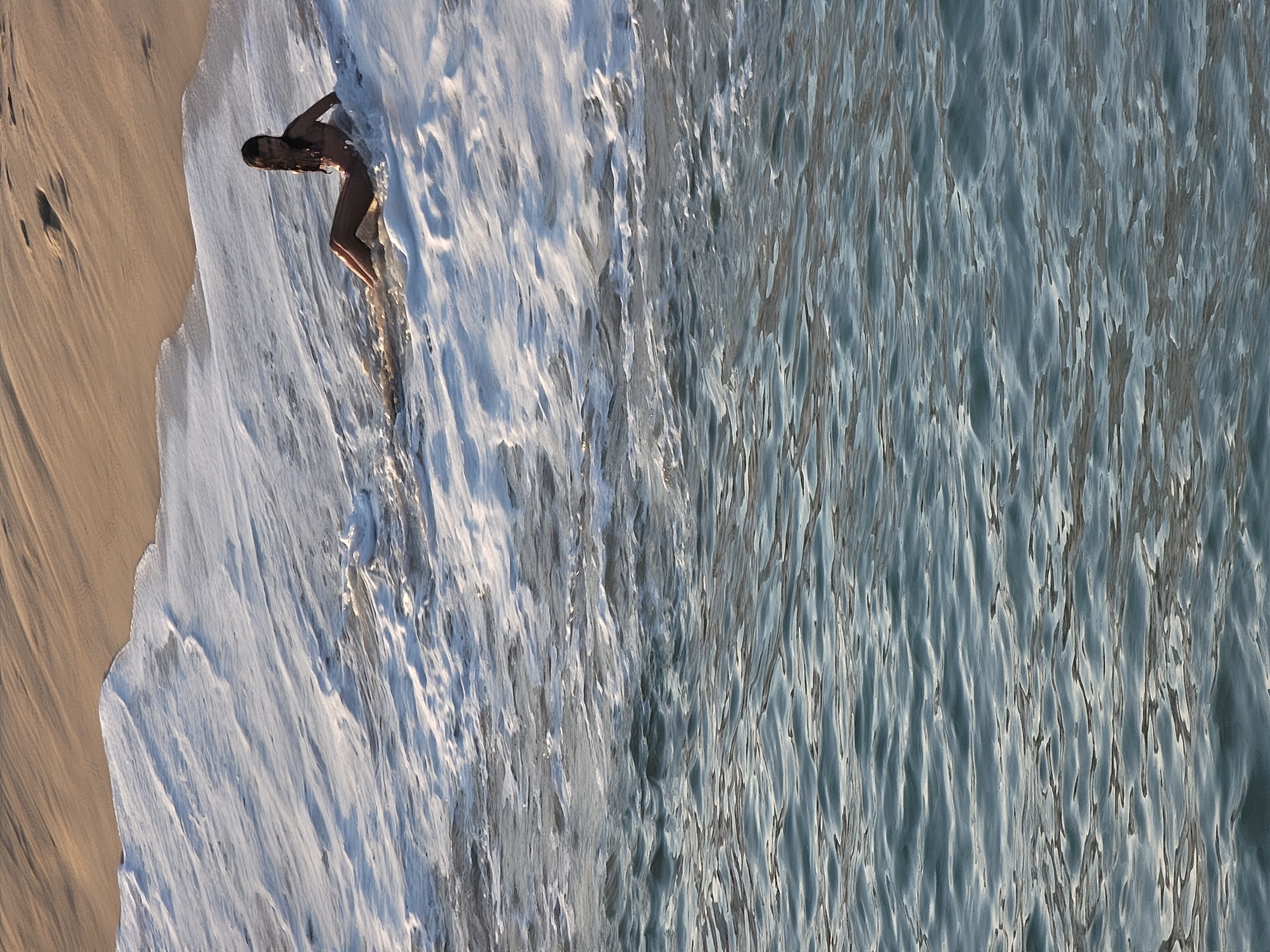 An aerial shot shows a young woman in a bikini sitting in the surf at a beach. She is facing to the right of the frame with her legs extended forward. The water around her is frothy white with breaking waves. The sand of the beach is visible in the upper right corner of the frame.