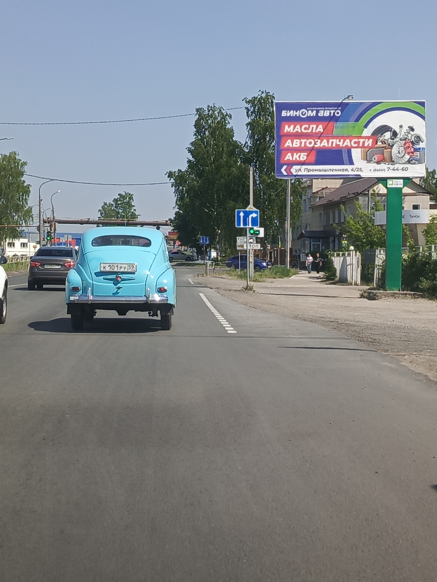 The image shows a light blue classic car with the license plate "K 101 PY 59" driving on a paved road. Behind it is a gray sedan. On the right side of the road is a large billboard advertising "BINOM AUTO" with the words "MASLA" (Oils), "AVTOZAPCHASTI" (Auto Parts), and "AKB" (Batteries). The address is given as "ul. Promyshlennaya, 4/25" which translates to Industrial street number 4/25, along with a phone number. A road sign with a blue arrow is also visible near the billboard. Two pedestrians are walking on the sidewalk. The location is the city of Chaikovsky, Russia.