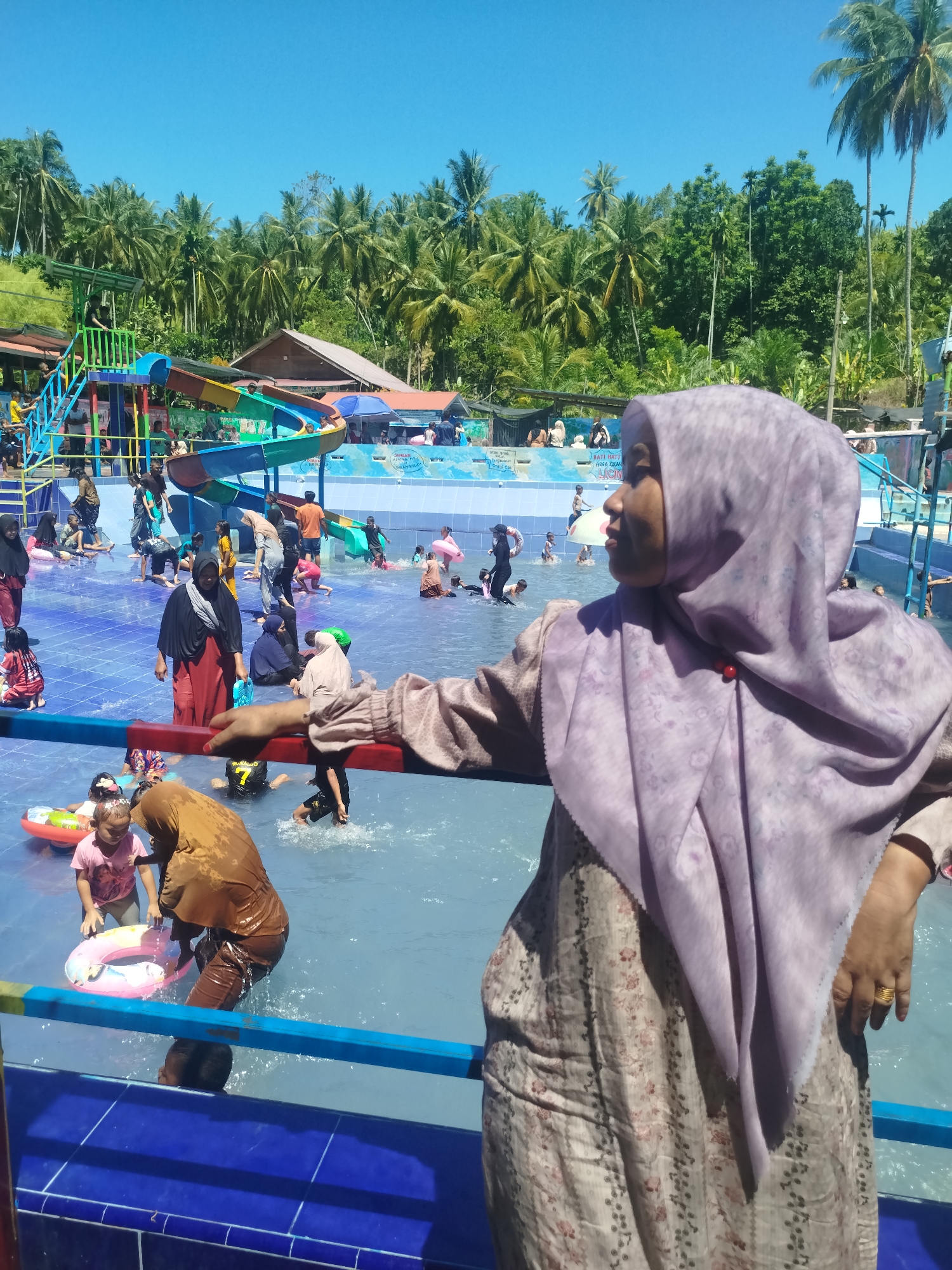 This is an outdoor image of a crowded water park in Palohseulimeng, Indonesia, on a bright, sunny day. In the foreground, a woman wearing a light purple hijab and a patterned dress leans against a blue railing. She is looking off to the right side of the frame. The middle ground features a pool filled with people of various ages wearing swimwear or casual clothes. Some are playing with inflatable rings. A water slide structure is visible in the background, as well as buildings, lush green trees, and a clear blue sky.
