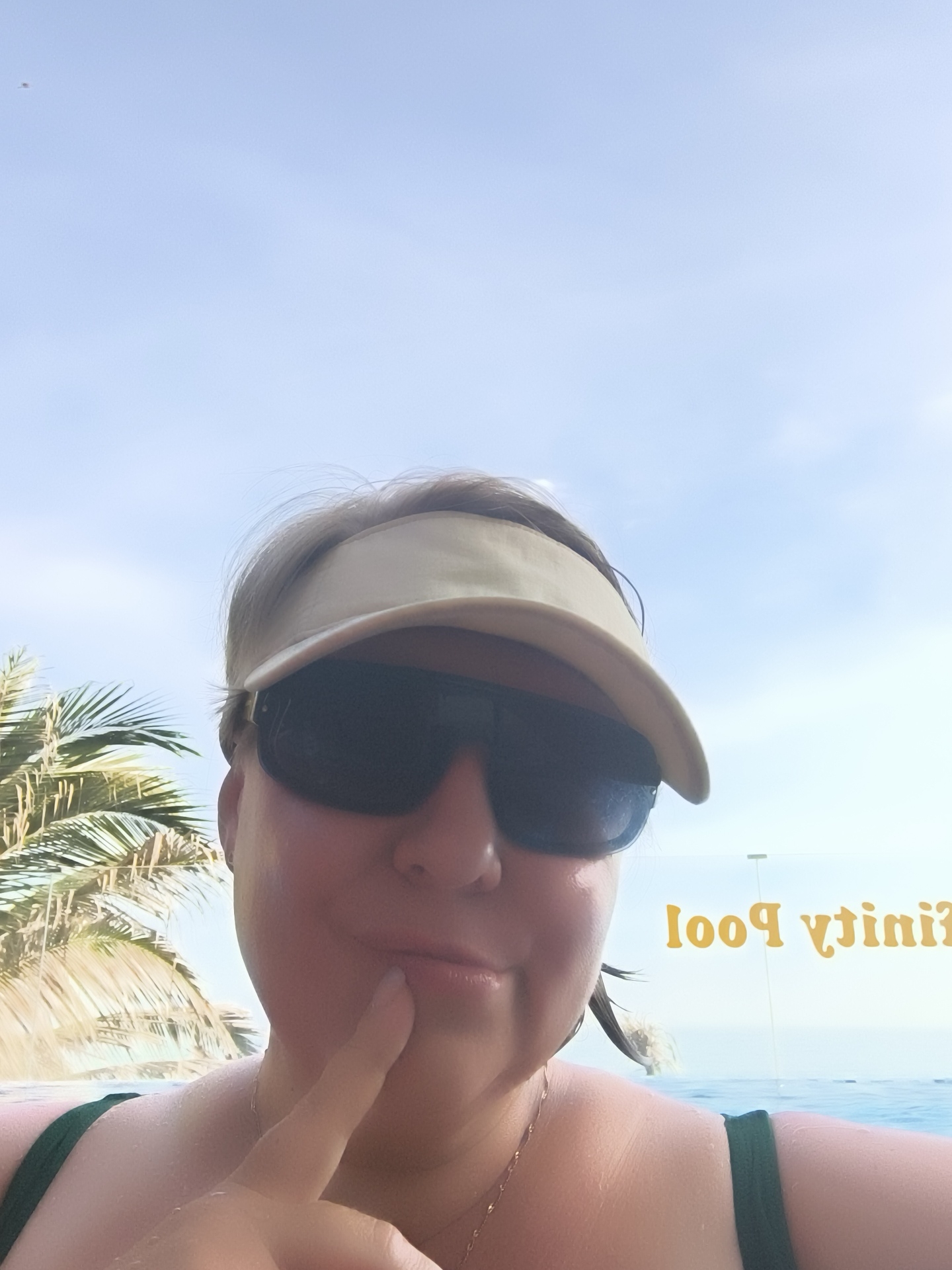 A woman is taking a selfie in what appears to be a swimming pool. She is wearing sunglasses and a light-colored visor. A palm tree is visible in the background to her left, and in the background to her right, text is visible on a glass barrier, appearing to read "Infinity Pool" backwards and upside down. The sky is bright blue with some wispy clouds. The woman has her finger to her chin, and she is wearing a green swimsuit.
