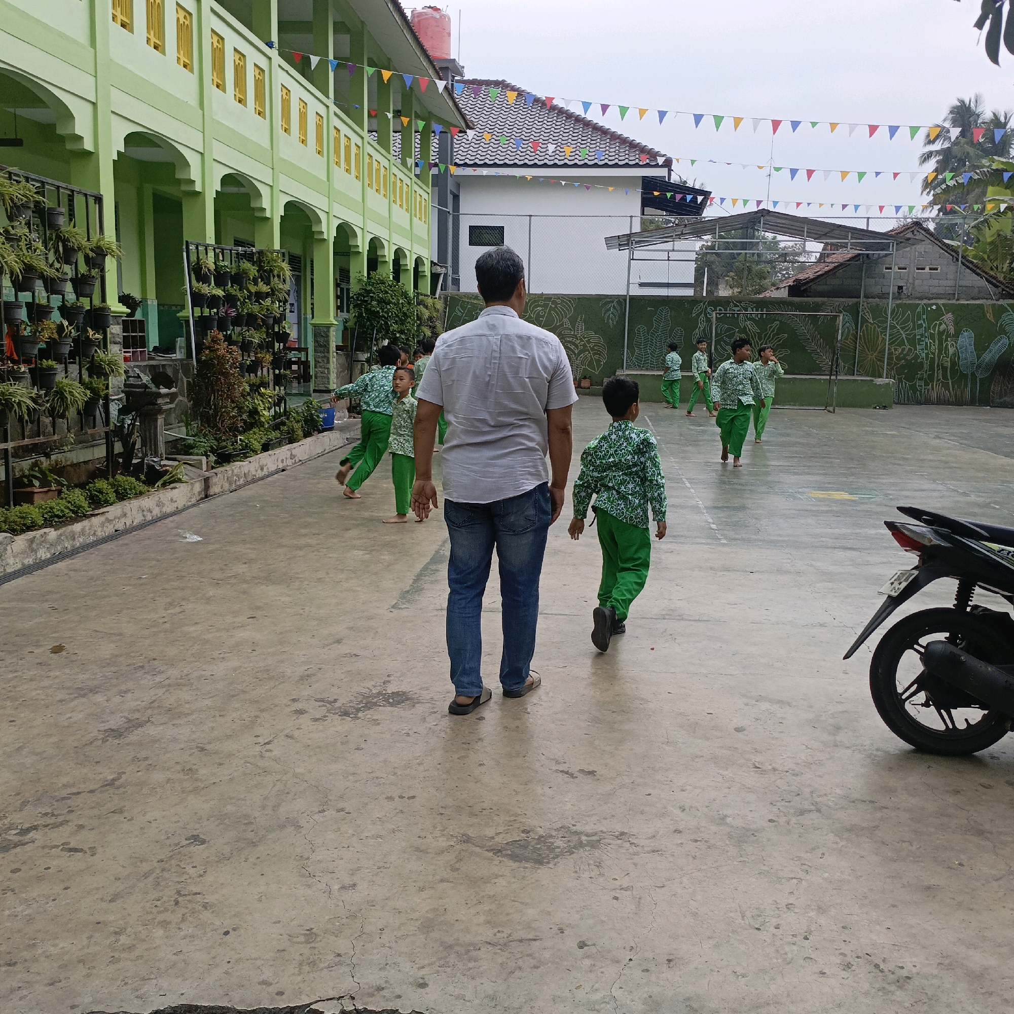 In Salatiga, Indonesia, a man in a grey shirt and blue jeans walks on a concrete courtyard, followed by a boy in a green patterned shirt and pants. Several other children, similarly dressed, are scattered across the courtyard, some in motion. Behind them, a light green building with yellow-trimmed windows stands tall, adorned with potted plants and hanging decorations. The scene appears to be an outdoor schoolyard, possibly during a recess or activity. A black motorcycle is parked on the right side of the image. The sky is overcast, suggesting a cloudy day.
