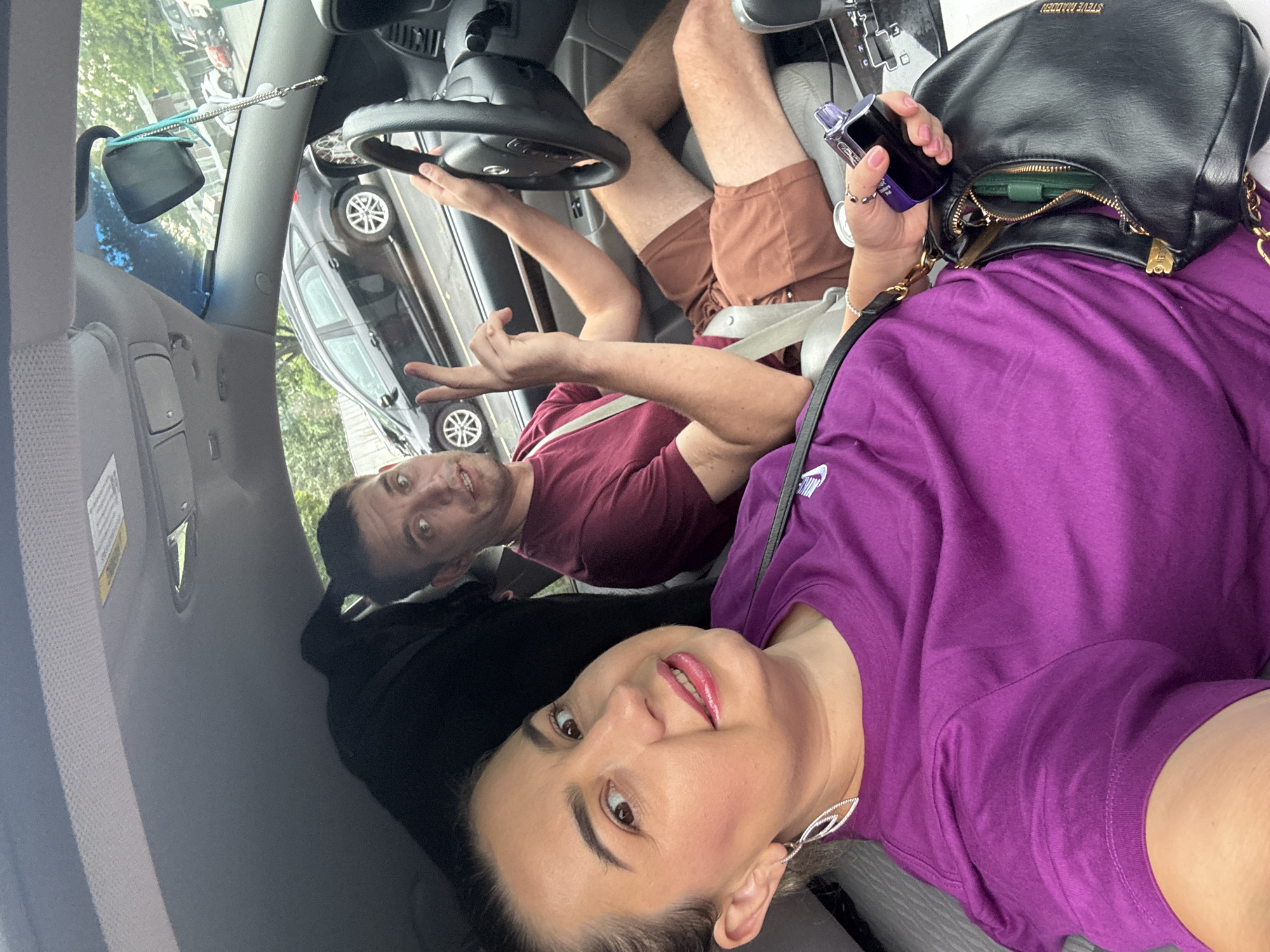 In a car, a couple is taking a selfie. The woman in the foreground is wearing a purple t-shirt and has her hair pulled back. She is smiling and looking at the camera. The man in the background is wearing a maroon t-shirt and is holding the steering wheel. He is making a peace sign with his fingers and looking at the camera. The car is in motion, as indicated by the blurry background. The car appears to be driving on a street in Wheeling, West Virginia, with other cars visible on the road and trees lining the street. The interior of the car is visible, including the dashboard, steering wheel, and seats. A black purse is visible in the woman's lap, and a bottle of perfume is in her hand.