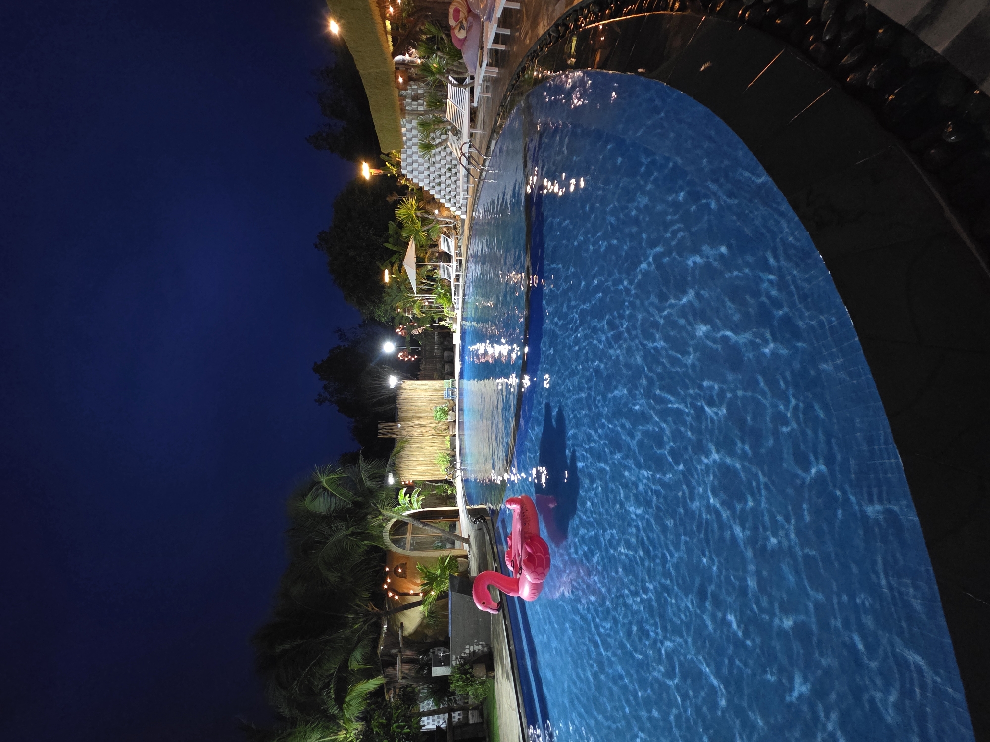 A nighttime photograph captures a swimming pool with a flamingo float against the backdrop of Mũi Né, Vietnam. The pool is illuminated, revealing its bright blue water, and the surrounding landscape is dotted with palm trees. The sky above is a dark blue, suggesting a clear night. Buildings and structures are visible in the background, providing context to the location within the city.