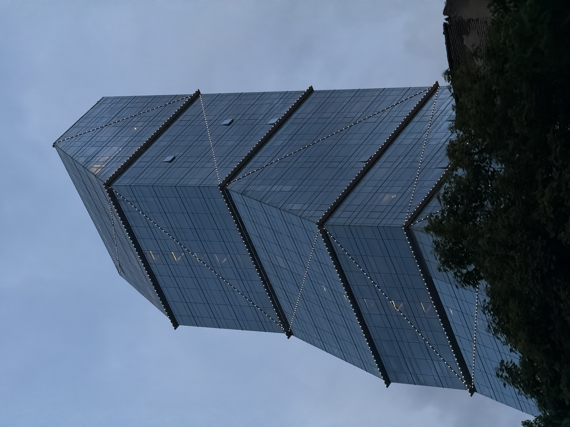 The image depicts the Axis Towers in Tbilisi, Georgia, from a low-angle perspective. The towers have a distinctive architectural design, with a faceted glass facade accented by strings of small, evenly spaced lights. The sky is overcast, suggesting dusk or dawn. In the right of the image, some trees block the view of the structure.