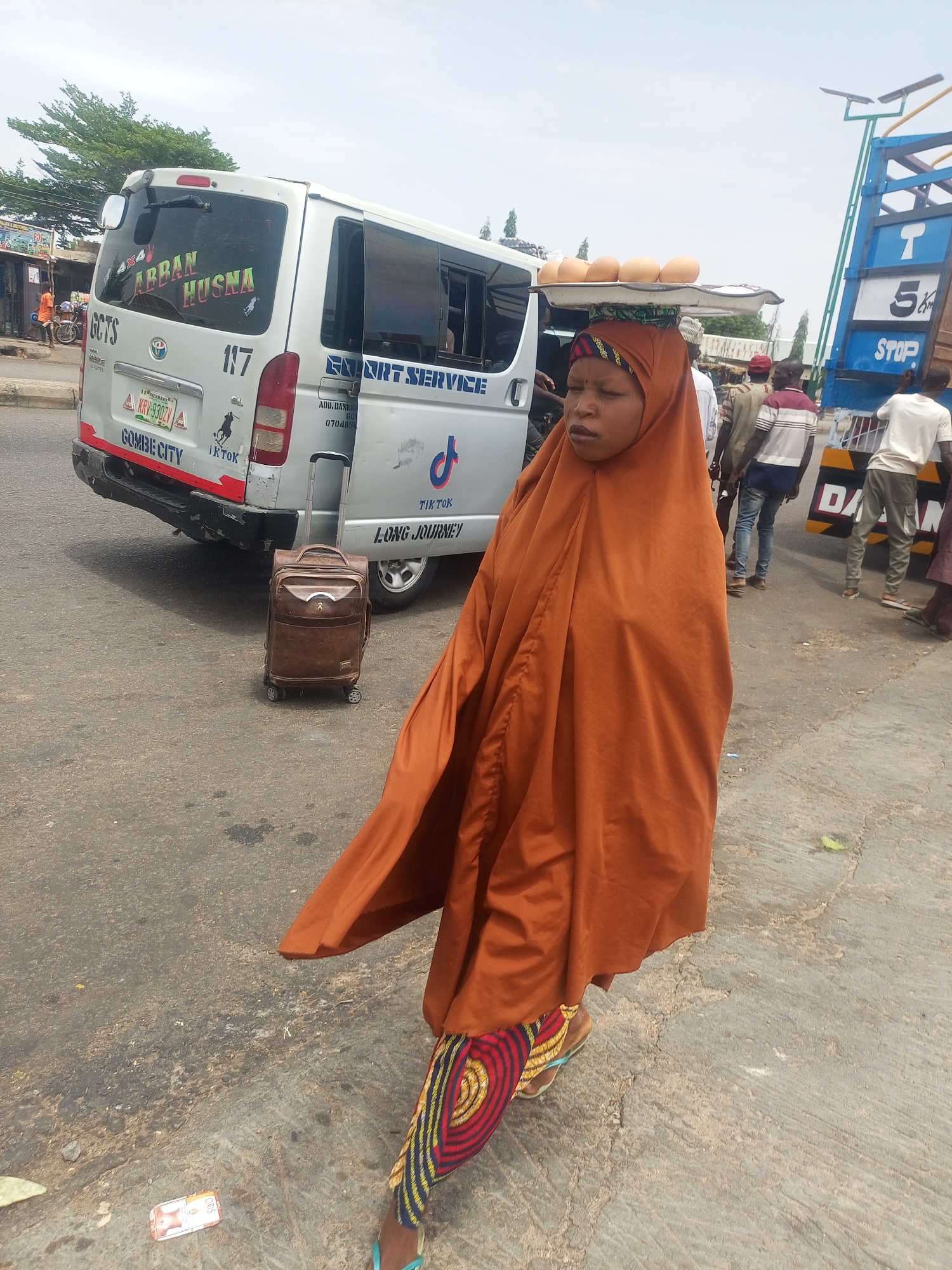 A woman in an orange hijab and patterned pants walks on a cracked asphalt street in Gombe City, Nigeria. She carries a tray of eggs on her head. Behind her is a white Toyota HiAce bus labeled "ABBAN HUSNA" and "GOMBE CITY", with the license plate KRV-930ZVA and markings for "EXPORT SERVICE" and "TIKTOK LONG JOURNEY." A brown suitcase stands next to the bus. Several people are standing near a truck with the word "STOP" on its side. Cigarette packaging is visible on the ground near the woman.