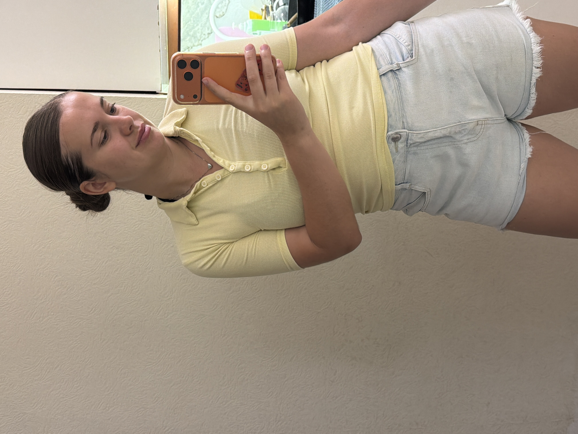 A young adult female is depicted taking a mirror selfie in an indoor setting, identified as being in the city of Sochi, Russia. She is positioned centrally, viewed from the upper torso to the upper thighs. Her dark brown hair is pulled back into a low bun, and she has a fair complexion, looking downward towards the phone she holds. She wears a light yellow, short-sleeved, ribbed polo shirt with a collared neckline and a five-button placket, with the top two buttons unfastened. A thin silver necklace with a small heart pendant is visible around her neck, along with a dark choker or band. She is wearing light wash denim shorts with frayed hems.
In her right hand, she holds an orange-cased smartphone featuring a triple-lens camera array on its rear, with a small object or sticker affixed to the back of the case. The background, visible in the mirror's reflection, consists primarily of a light beige wall with a subtle, textured, swirling pattern. To the subject's right, a vertical white panel is visible. Further right, a partial view of what appears to be an illuminated aquatic or terrarium setup, containing water and some green elements, is present. A dark countertop or surface, along with various bottles including a green pump dispenser, are also visible in the reflection to the right of her waist area. The lighting is even, consistent with an indoor environment.