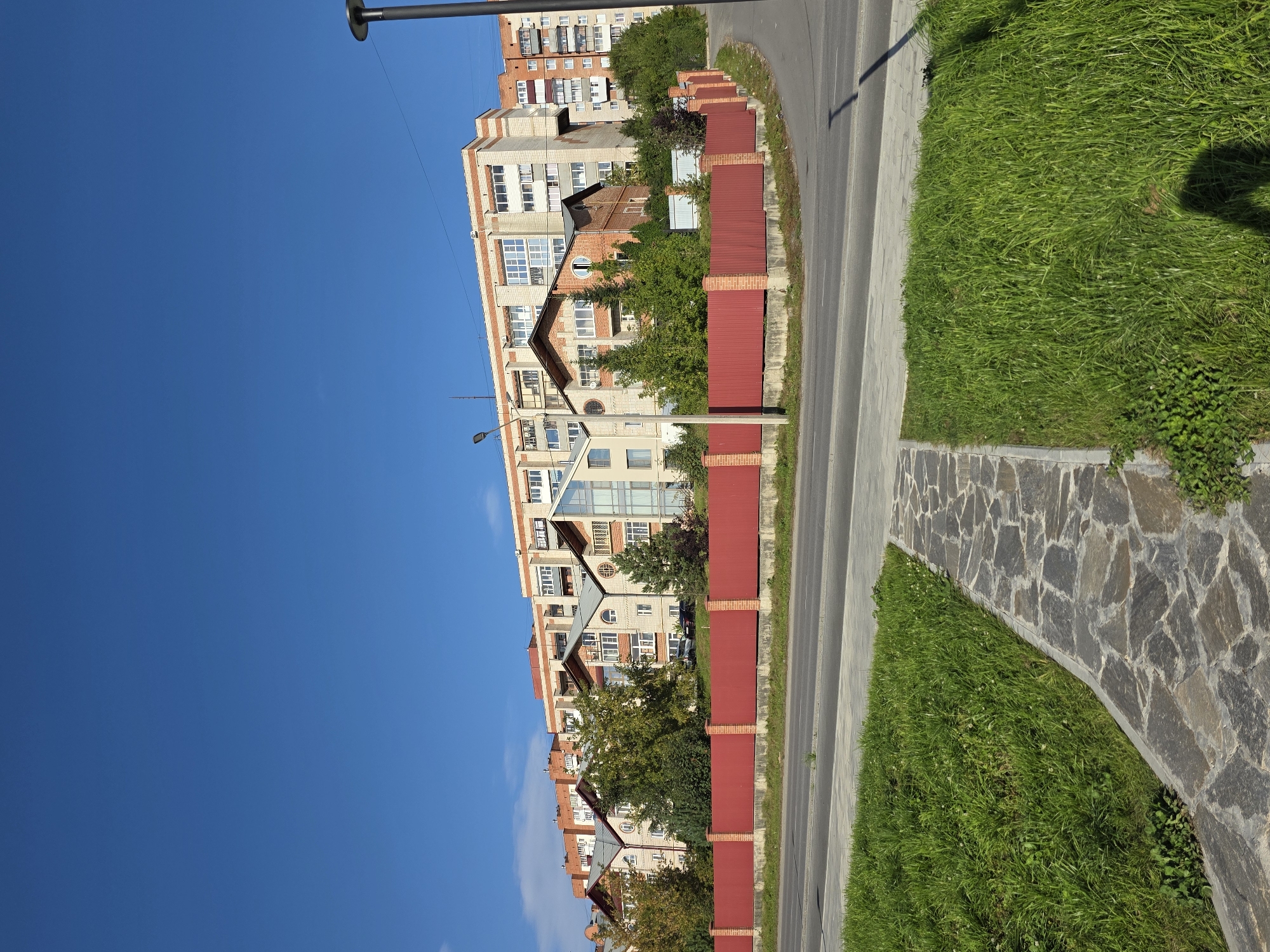 The image depicts a residential area in Miass, Russia, on a clear day under a bright blue sky. In the foreground, a paved walkway made of irregular grey stones is visible, bordering a lush green lawn. Beyond the lawn lies a red metal fence supported by brick posts, separating the walkway and lawn from a street. The street itself is paved with asphalt and appears to be a typical urban road.
Behind the red fence, a row of multi-story apartment buildings is visible. These buildings exhibit a mix of architectural styles, with some featuring brick exteriors and others appearing to be clad in lighter-colored materials. Many windows are visible, indicating occupied residences. Green trees and shrubs are interspersed among the buildings, adding a touch of nature to the urban landscape. A street lamp is present on the right side of the image, casting a shadow on the ground. The overall scene suggests a well-maintained urban environment. There are no people visible in the image.