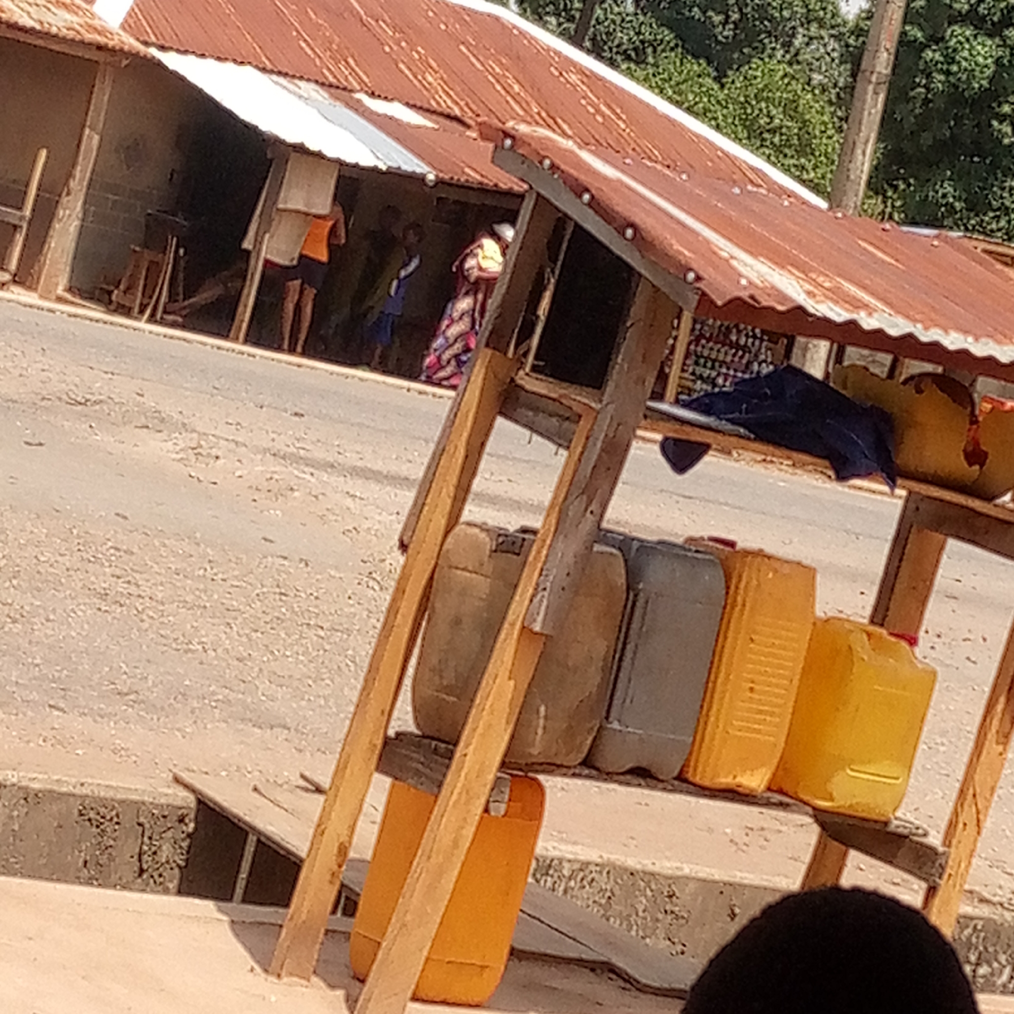 This image depicts an outdoor scene in an unspecified urban location. In the foreground, a makeshift wooden structure is visible, laden with several plastic containers, some of which are yellow and one is gray. A dark-colored cloth is draped over the top shelf. The wooden structure appears to be some sort of display or stand.
Beyond the structure, a road stretches into the distance, alongside a building with a corrugated metal roof that is partly rusted. Several individuals are present near the building; one is wearing an orange top and dark shorts, while another is wearing a colorful patterned garment. Other figures are somewhat obscured within the building's shaded area. The general environment seems to be a neighborhood or roadside area.