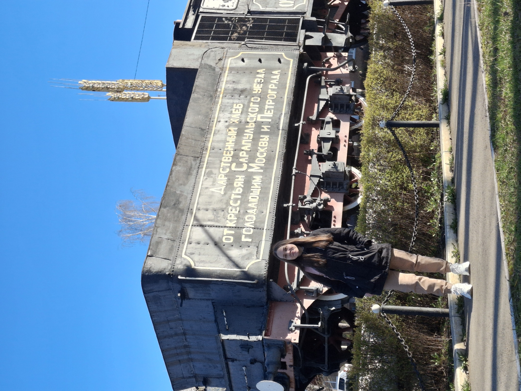A full shot depicts a young woman standing next to a train in Sarapul, Russia. The woman is wearing a black jacket, tan pants, and white sneakers. She is positioned on a sidewalk next to the train, and she is facing the camera. The train is large and black, with red wheels. It is decorated with gold accents. Text on the side of the train is in Russian. The train is behind a black metal chain barrier. The sky is blue and cloudless.