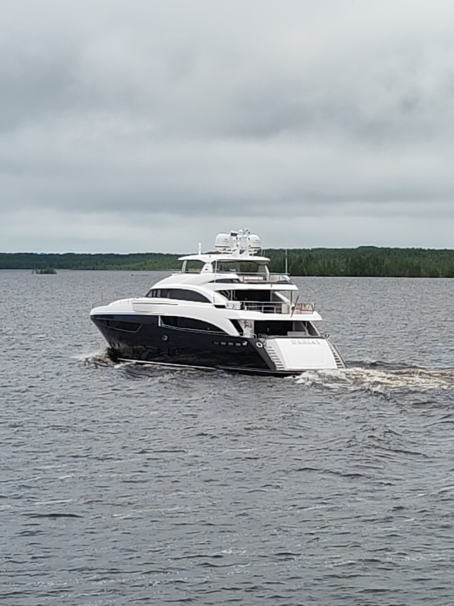 The image shows a large, modern yacht cruising on a body of water, likely a lake or wide river. The yacht has a dark blue and white hull with multiple decks and a radar or satellite dome on top. The name "DARIA" is visible on the stern. The water is choppy with the wake of the yacht clearly visible. The shoreline in the background is lined with trees and vegetation. The sky is overcast. No people are clearly visible in the image. The location is likely near a forested area with a body of water. The name "Gorushka, Russia" may be relevant, but cannot be confirmed with the information present in the image.