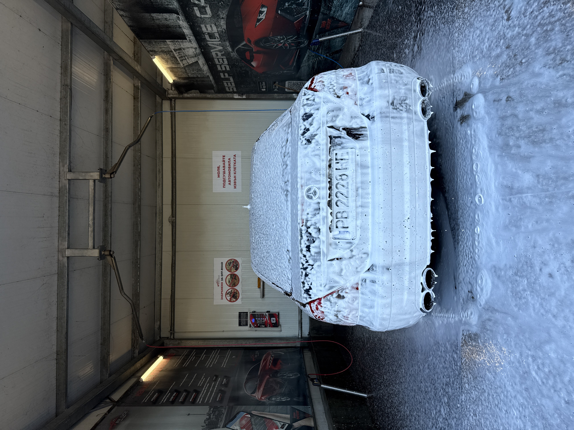 The image shows a car covered in white foam at a self-service car wash. The location is identified as Sofia, Bulgaria, based on the Bulgarian license plate "PB 2228 HE" on the car. The car is being washed with a pressure washer, the wand of which is visible near the bottom of the frame. The car is inside a car wash bay, as indicated by the overhead supports and advertisements on the walls. Signage in Bulgarian is visible, including instructions to dry the car outside the bay and a warning against off-road vehicles. There is an advertisement for "SELF SERVICE CAR" and an image of a red sports car on the wall. The ground around the car is wet and covered with foam residue.