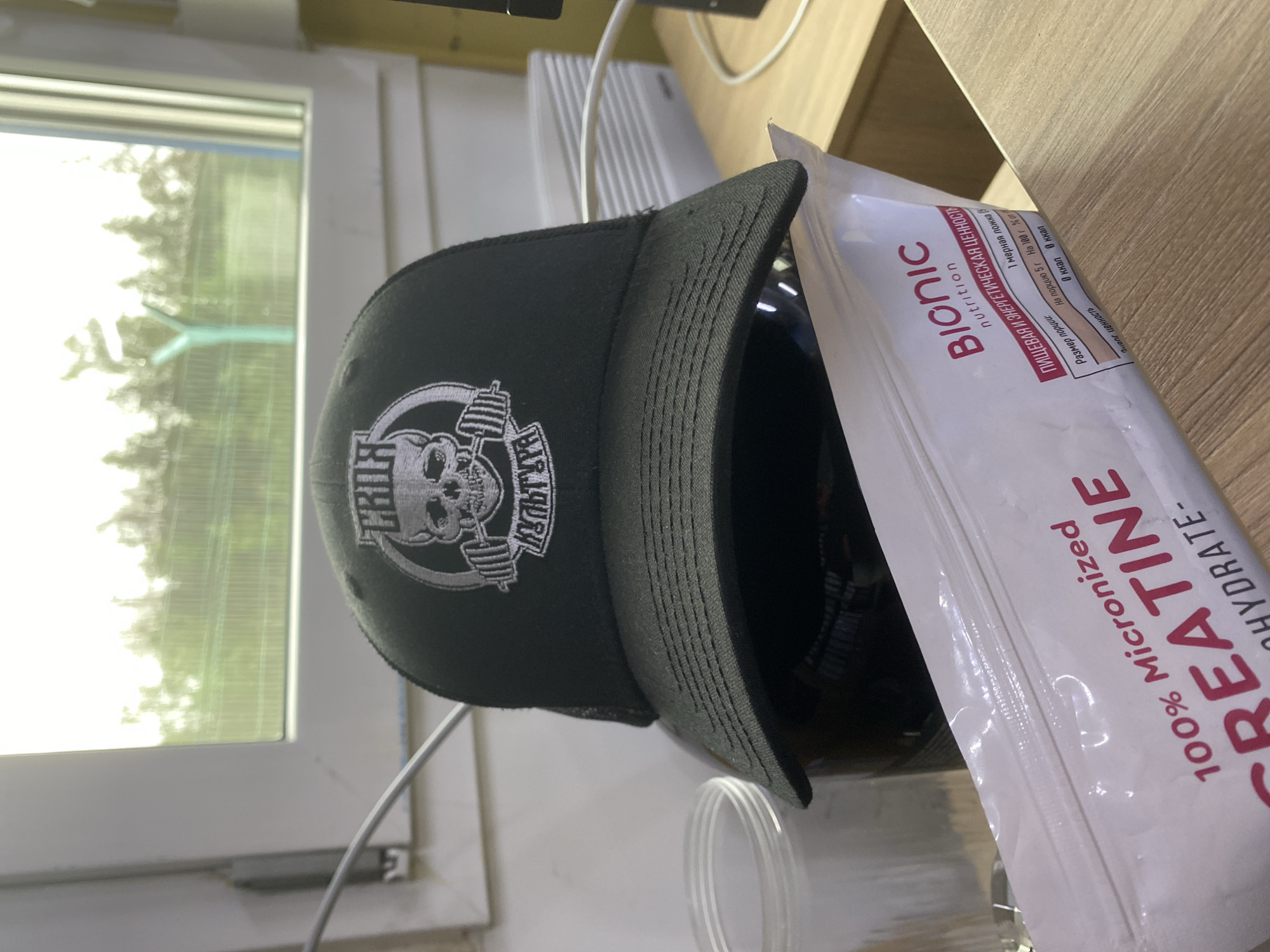 The image depicts a black trucker hat with a white embroidered logo of a skull holding a barbell and the Cyrillic text "ТВОЯ КУЛЬТУРА". The hat is placed on top of a black helmet. In the foreground, a white resealable pouch of "100% Micronized CREATINE" from "Bionic Nutrition" is visible. The pouch has Russian text indicating nutritional information. To the left, a clear plastic container with a ribbed texture is partially visible. The background shows a window with a view of trees and a pale green structure, possibly a fence or part of a building, against a bright sky. The scene appears to be indoors, with a wooden surface in the foreground and white, ribbed objects resembling electronics or ventilation in the background. The lighting suggests it is daytime. There are no people visible in the image. The location of Luzhitsy, Russia, is not explicitly indicated by any visual cues in the image.