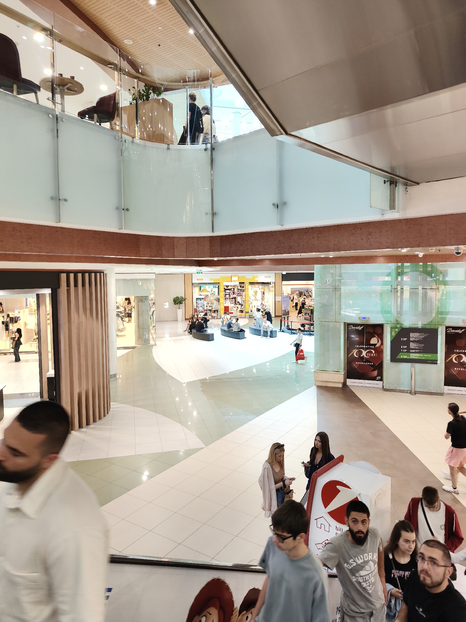 This image shows an indoor public space, likely a shopping mall, with several levels and a central atrium. There are people in the foreground, middle ground, and background. In the foreground, a man in a white collared shirt is blurred and walking towards the camera. Behind him, other individuals are visible, some walking down an escalator.
The middle ground features a wide, tiled walkway with multiple shops and seating areas. People are seen walking, sitting, and interacting in these areas. A prominent architectural feature is a glass-paneled balcony overlooking the atrium.
In the background, more shops are visible, along with decorative elements and lighting. The overall environment appears clean and modern, with polished floors and contemporary design.
The provided context indicates this scene is located in the city of Sofia, Bulgaria. Without further information, it is not possible to identify the specific shopping mall.