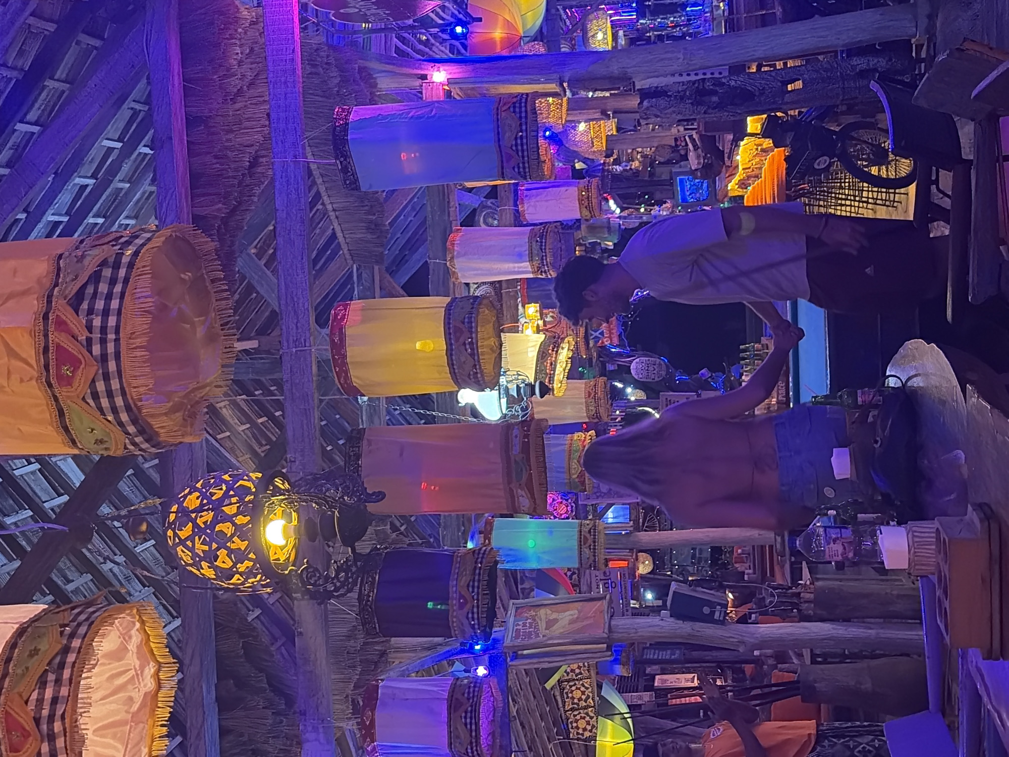 The image depicts a lively bar scene at night in Banjar, Indonesia. The atmosphere is illuminated by colorful, hanging lanterns of various shapes and sizes, casting a vibrant glow. In the foreground, a couple stands by a bar, holding hands, with the woman facing away from the camera and the man facing towards it. They appear relaxed and engaged with each other. Behind them, the bar area is visible, with bottles, drinks, and other bar paraphernalia. A few other individuals are present in the background, suggesting a social and bustling environment. The architecture of the bar incorporates natural materials like wood and thatch, adding to the tropical ambiance. The overall impression is one of a vibrant and engaging night out in a tropical setting.