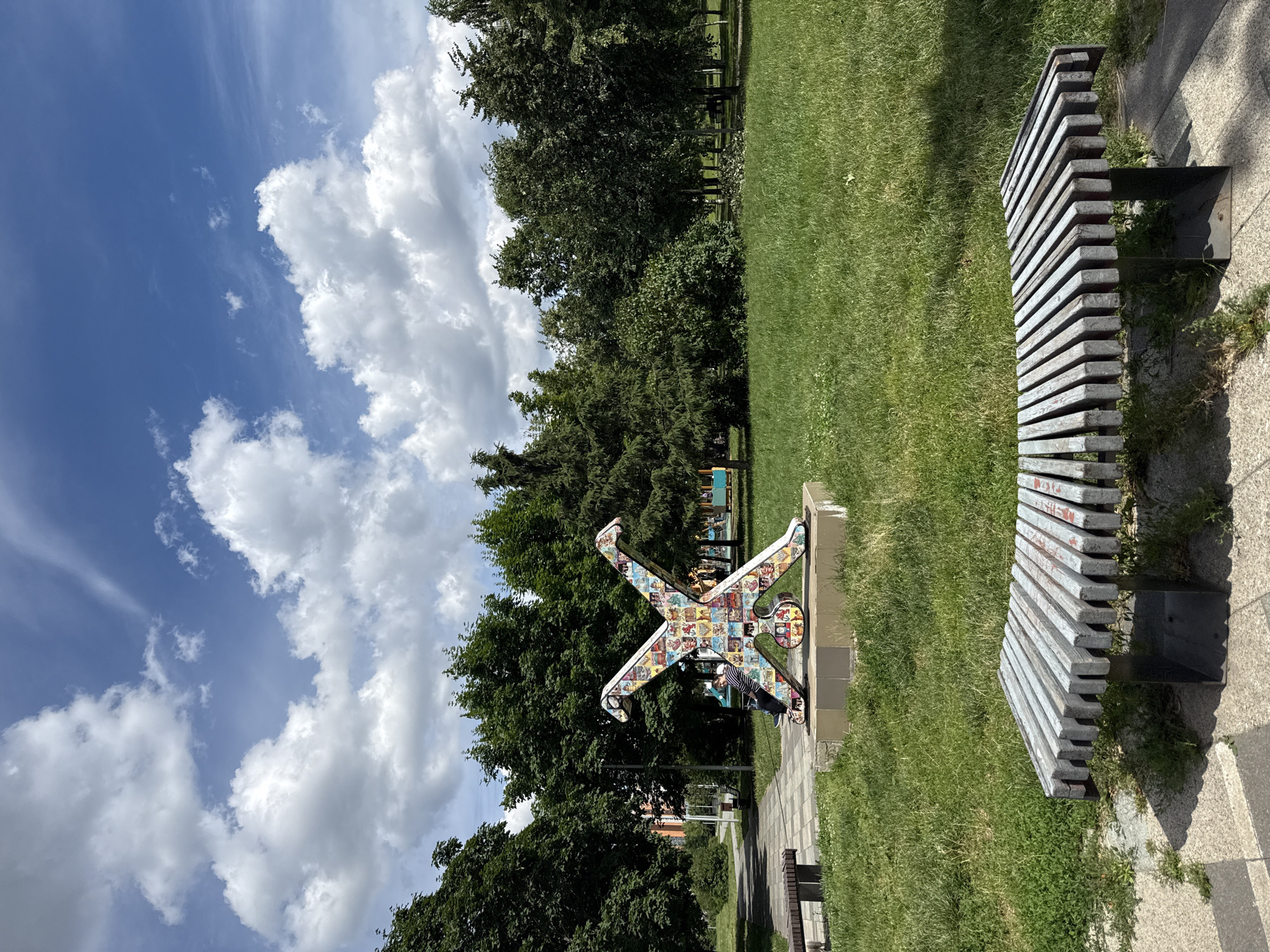The image shows an outdoor scene in Moscow, Russia, on a sunny day with scattered clouds. The foreground features a modern park bench with a weathered, slatted design. To the left of the bench, a vibrant, abstract sculpture resembling a person in a cartwheel pose stands on a concrete base. The sculpture is decorated with colorful, mosaic-like panels.
In the background, a lush green lawn stretches towards a line of dense trees, suggesting a park or recreational area. Beyond the trees, glimpses of buildings are visible, indicating an urban setting. The sky is a bright blue, with dramatic white cumulus clouds casting shadows on the grassy areas. The lighting suggests it is midday. There is no discernible text in the image.