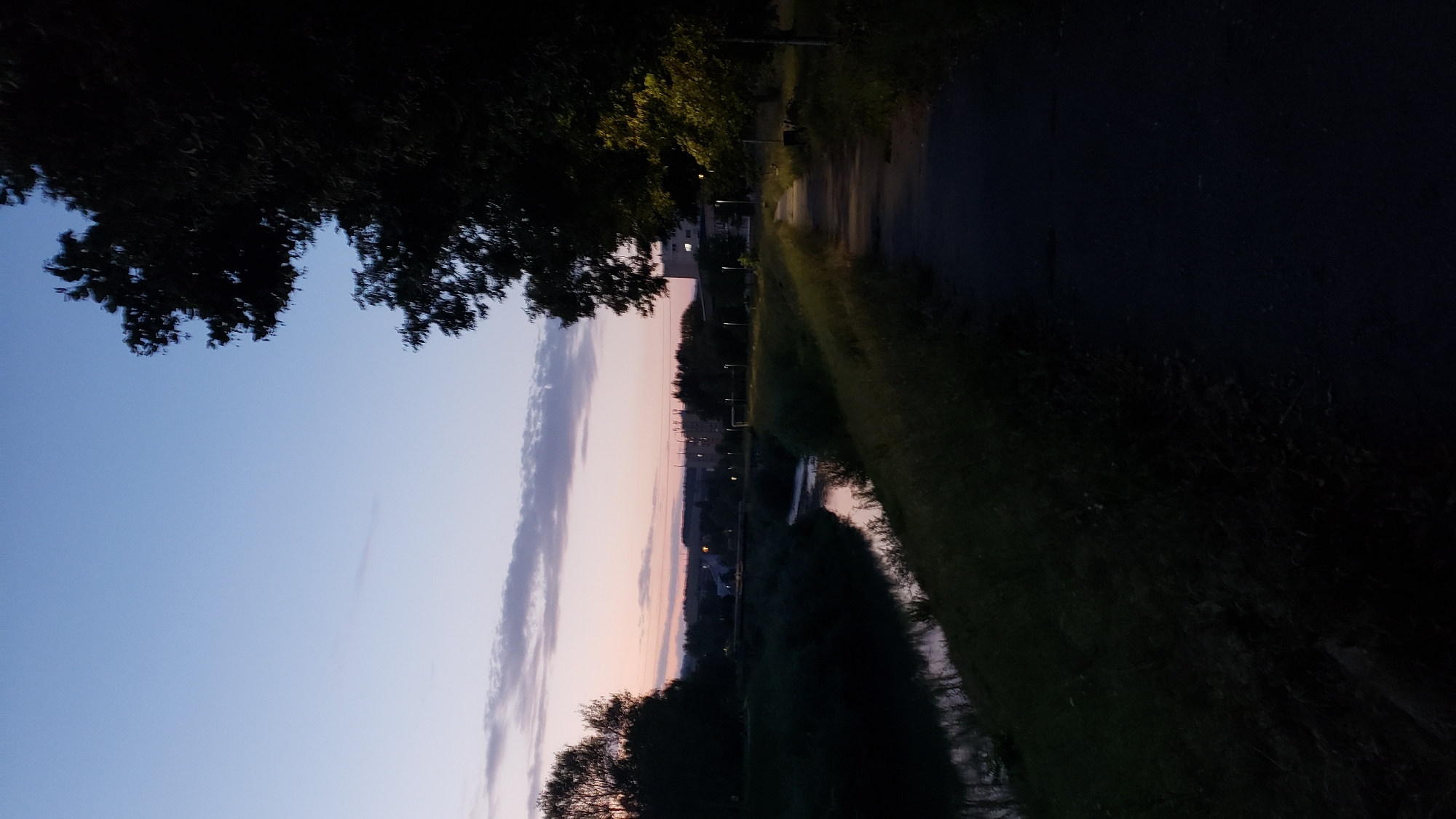 The image depicts a tranquil evening scene along a riverbank in Prešov, Slovakia. A paved pathway runs along the right side of the frame, bordered by lush green grass and trees. The river meanders through the left side of the image, reflecting the soft hues of the twilight sky. In the distance, buildings and industrial structures are visible, silhouetted against the horizon. The sky is a gradient of soft blues and pinks, with scattered clouds. The overall atmosphere is serene and peaceful, suggesting a quiet moment in the city.