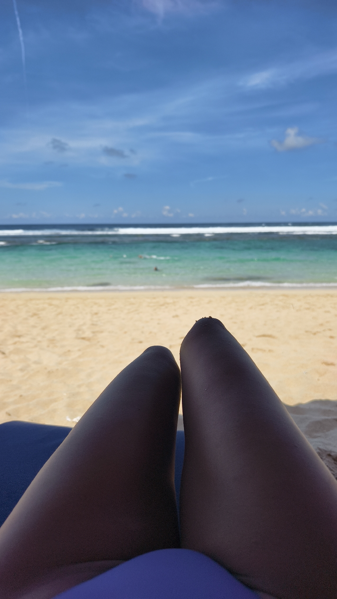 The image shows a first-person perspective of someone relaxing on a beach in Ungasan, Indonesia. The person is sitting in a blue beach chair, and their dark-skinned thighs are visible in the foreground. The beach stretches out in front of them, with tan sand leading to turquoise water and white waves. In the distance, the ocean meets the sky under a blue sky dotted with scattered clouds. There are a few people visible in the water.