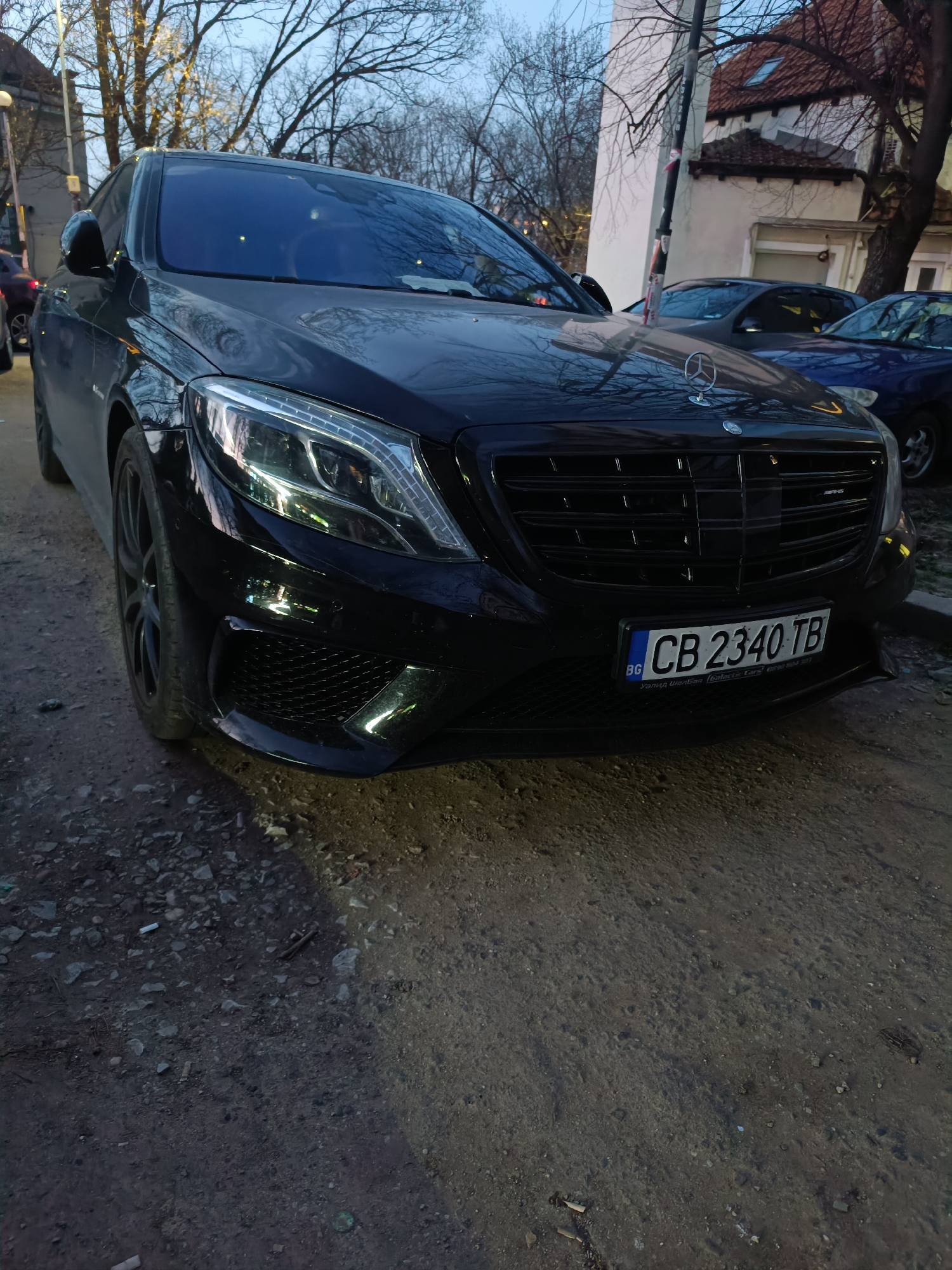 The image shows a black Mercedes-Benz S-Class, likely an AMG variant, parked on a dirt and gravel surface. The license plate reads "CB 2340 TB" and indicates registration in Bulgaria (BG). The vehicle is positioned on a street with other cars parked nearby and buildings visible in the background. Bare trees suggest either late autumn or early spring. The scene likely takes place in a neighborhood on the outskirts of Sofia, Bulgaria, given the visible architecture and general environment. The time of day appears to be late afternoon or early evening, judging by the lighting conditions.