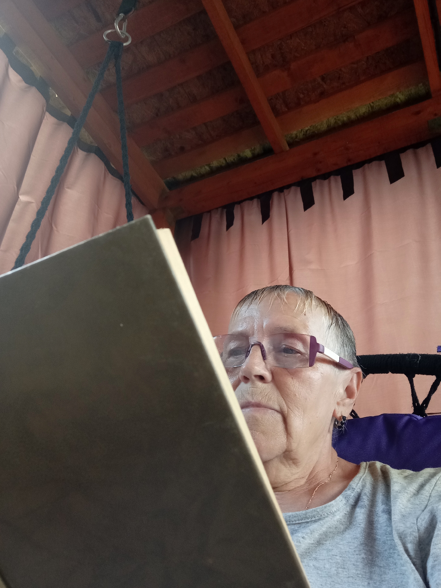 An elderly woman with short, graying hair and purple-rimmed glasses is seated, holding a large book in her hands. She is wearing a light gray t-shirt and a delicate gold chain necklace. The woman appears to be reading the book, her gaze directed downwards. She is sitting on a purple cushion, possibly part of a chair or swing, within an outdoor structure with a wooden roof and pink curtains. The background shows the textured wooden beams of the roof. The scene is captured from a low-angle perspective, looking up towards the woman and the structure's interior. The location is identified as Degtyarsk, Russia.