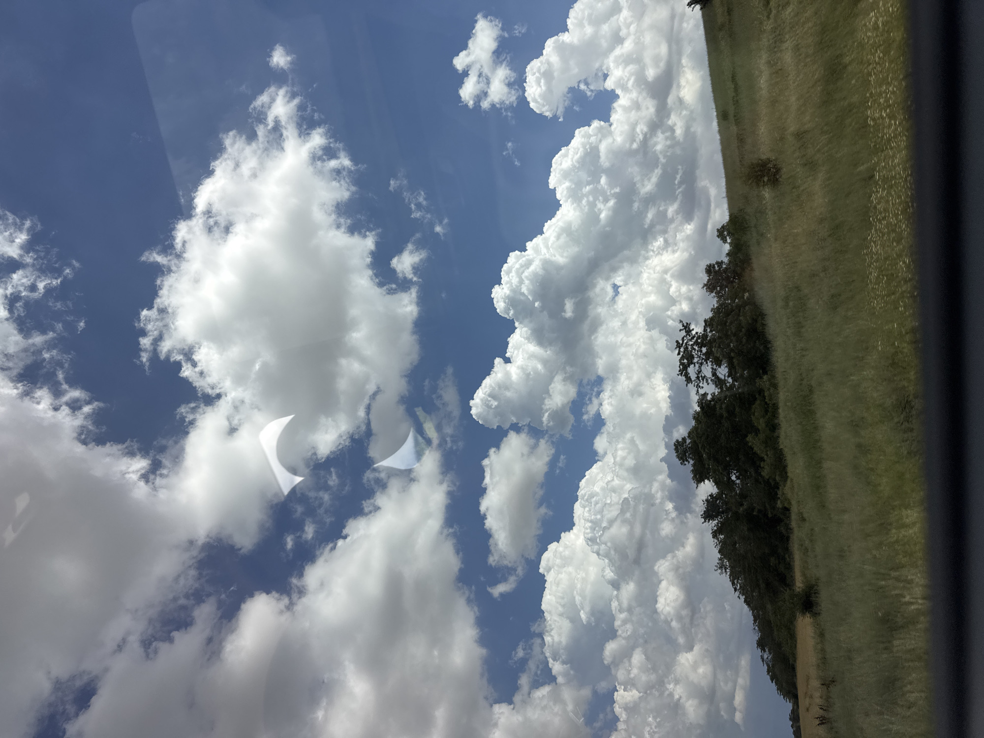 The image depicts a bright, cloudy sky with a landscape of green fields and trees. The sky takes up a significant portion of the frame, filled with fluffy white cumulus clouds against a vivid blue backdrop. There are two distinct reflections on what appears to be a window or glass surface, one of which is a bright, crescent-shaped white reflection.
The landscape on the right side of the image is a rolling green field, appearing slightly dry and tall with scattered lighter-colored vegetation, possibly wildflowers. A line of dark green trees borders the field, suggesting a wooded area or forest edge. The lighting indicates it is daytime, with the sun casting bright light on the clouds and landscape. The weather appears to be fair, with dramatic cloud formations suggesting potential for passing showers or just a dynamic sky.
The image is oriented sideways, with the landscape running diagonally from the bottom left to the top right. A dark, out-of-focus object, likely part of a vehicle or frame, is visible along the right edge of the image. No people or specific objects are clearly identifiable, beyond the natural elements of the sky and landscape. The overall mood is serene and expansive, capturing a moment of natural beauty. There is no visible text in the image.