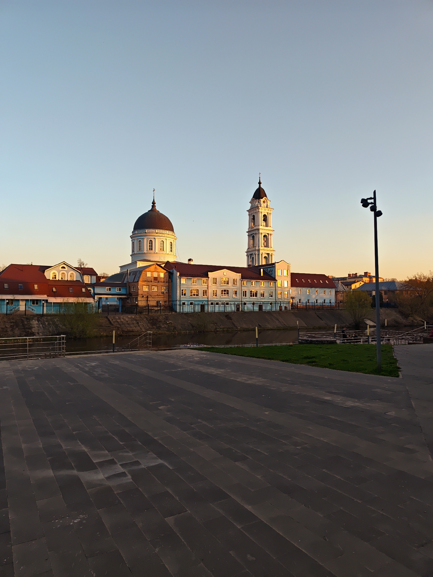 The image captures a view of the cityscape of Noginsk, Russia at dusk or dawn, with the sky gradienting from light blue to warm orange hues. In the foreground, a gray-paved square leads the eye towards a body of water, possibly a river, bordered by a concrete embankment and metal railings. Beyond the water, a row of buildings stands, consisting of a light-colored church with a prominent dark dome and a tall, white bell tower. Adjacent to these religious structures are buildings with red roofs and blue facades, some appearing to be residential or commercial. A black lamppost is located on the right side of the frame. There are no people visible in the image. The scene appears calm and peaceful, devoid of motion or activity.