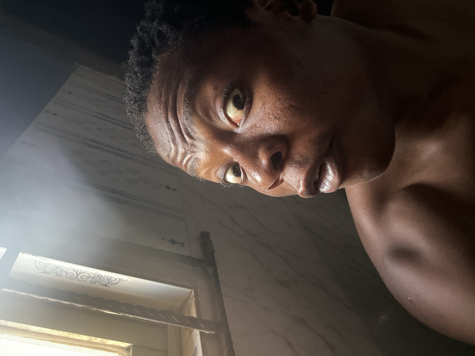 A close-up, high-angle portrait shows a dark-skinned person with short, dark hair. The person is facing the camera with their head tilted to the right. Their eyes are wide and expressive. The person appears to be wet, with a glistening complexion. The background is out of focus but suggests a room with a window. The location is Unknown, Unknown. The environment suggests a confined space.