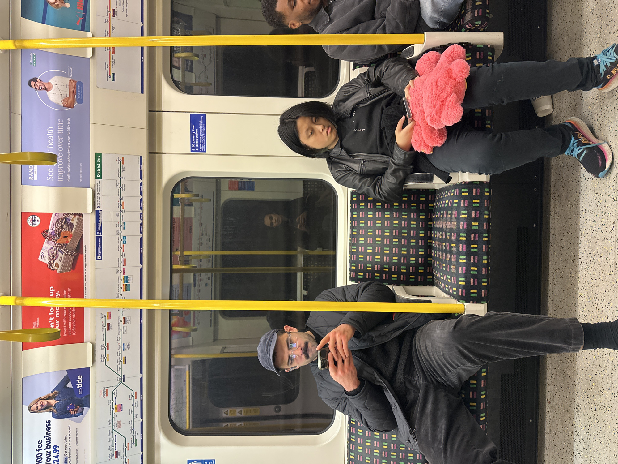 The image depicts the interior of a London Underground train carriage. Three individuals are visible: one man in a black jacket and cap is seated, looking at his phone; a woman in a black leather jacket holding a pink plush item is seated with her eyes closed; and the top of another man's head is visible in the background. The train seats have a multicolored patterned upholstery. Yellow handrails run horizontally across the frame. Advertisements are visible on the carriage walls, promoting services like RAND Health and Tide business accounts, along with route maps of the District Line. A reflection is visible in the window behind the woman.