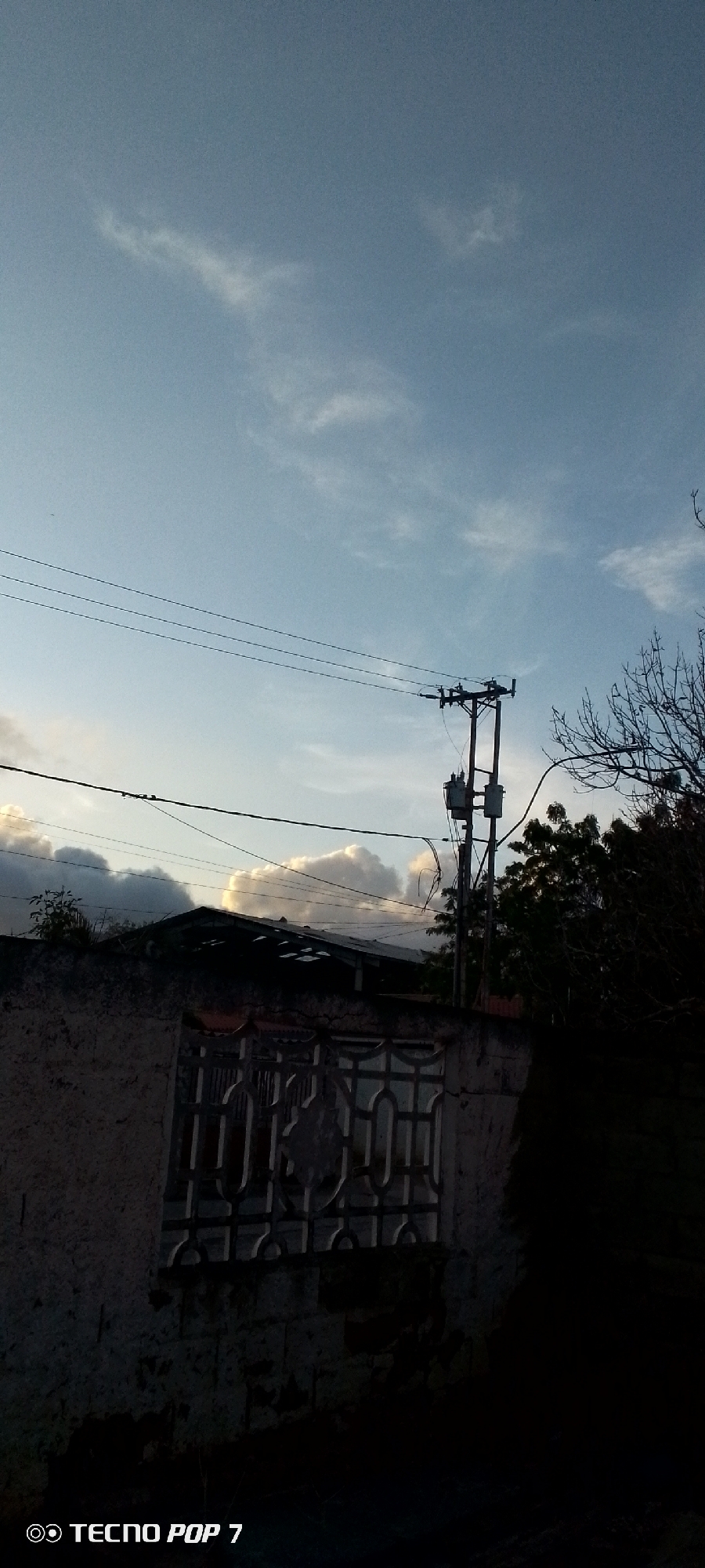 The image captures a partially obscured view of a building exterior in what appears to be a residential area. The sky is visible, with a mix of clear blue and some cloud formations. Utility lines and a utility pole with transformers are present against the sky. A portion of the building is visible, including a wall with a decorative window or gate feature. There are trees and other foliage in the background. The lower portion of the image is heavily shaded, rendering details unclear. The camera watermark indicates that the image was taken with a TECNO POP 7. While Altagracia, Venezuela, is specified as the location, there are no details within the image to confirm this location directly.