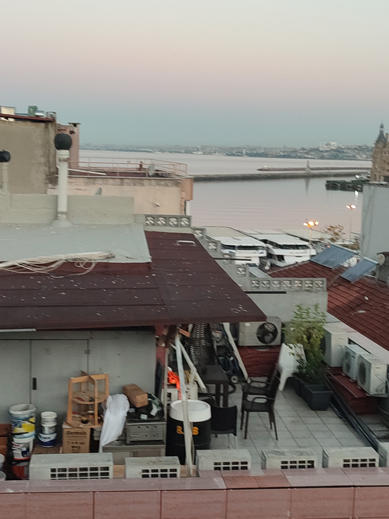 The image shows a view of rooftops in Kadıköy, Turkey, overlooking a body of water. In the foreground, there are several rooftops covered with dark red tiles, some with white speckles. Several air conditioning units are visible on one of the rooftops, along with outdoor furniture such as chairs and tables. Storage items, including buckets, boxes, and a wooden shelving unit, are present on a lower rooftop. In the mid-ground, two white passenger boats are docked, and streetlights with orange illumination are present. The background features a hazy city skyline with several buildings, including a prominent domed structure. The water body appears calm, and a distant lighthouse is visible. The sky is a soft gradient of pink and blue, suggesting either dawn or dusk. No people are visible in the frame.