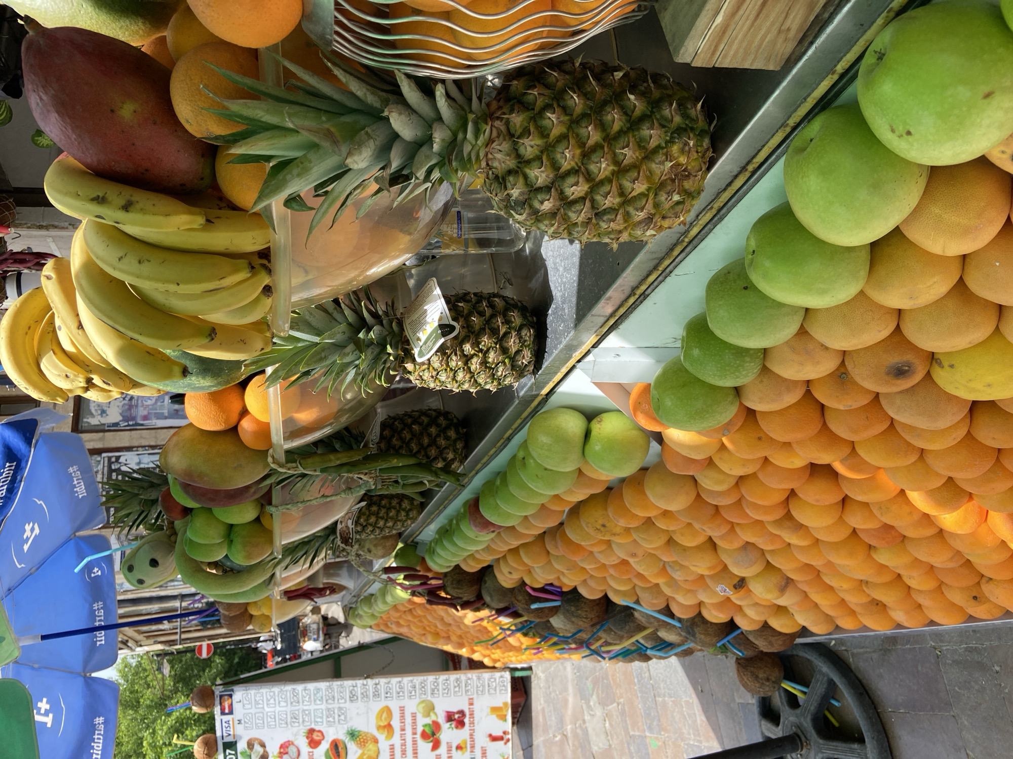 The image shows a fruit stand in Antalya, Turkey. The stand is stocked with a variety of fresh fruits, including bananas, mangoes, pineapples, oranges, grapefruits, green apples, and coconuts. There is also a menu listing different milkshake and fruit juice options. A blue umbrella with Turkish writing provides shade. The stand is positioned on a stone walkway. People are visible in the background.