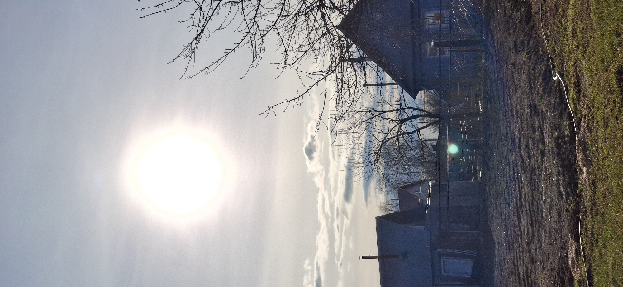 The image shows a rural scene, possibly in the vicinity of Chaikovsky, Russia. The sun shines brightly in the sky, partially obscured by thin clouds. Bare tree branches reach into the upper portion of the frame, silhouetted against the light. The roofs of several buildings are visible, suggesting a small village or residential area. A strip of land transitions from bare soil to grass on the right side of the image. The image is vertically oriented.
